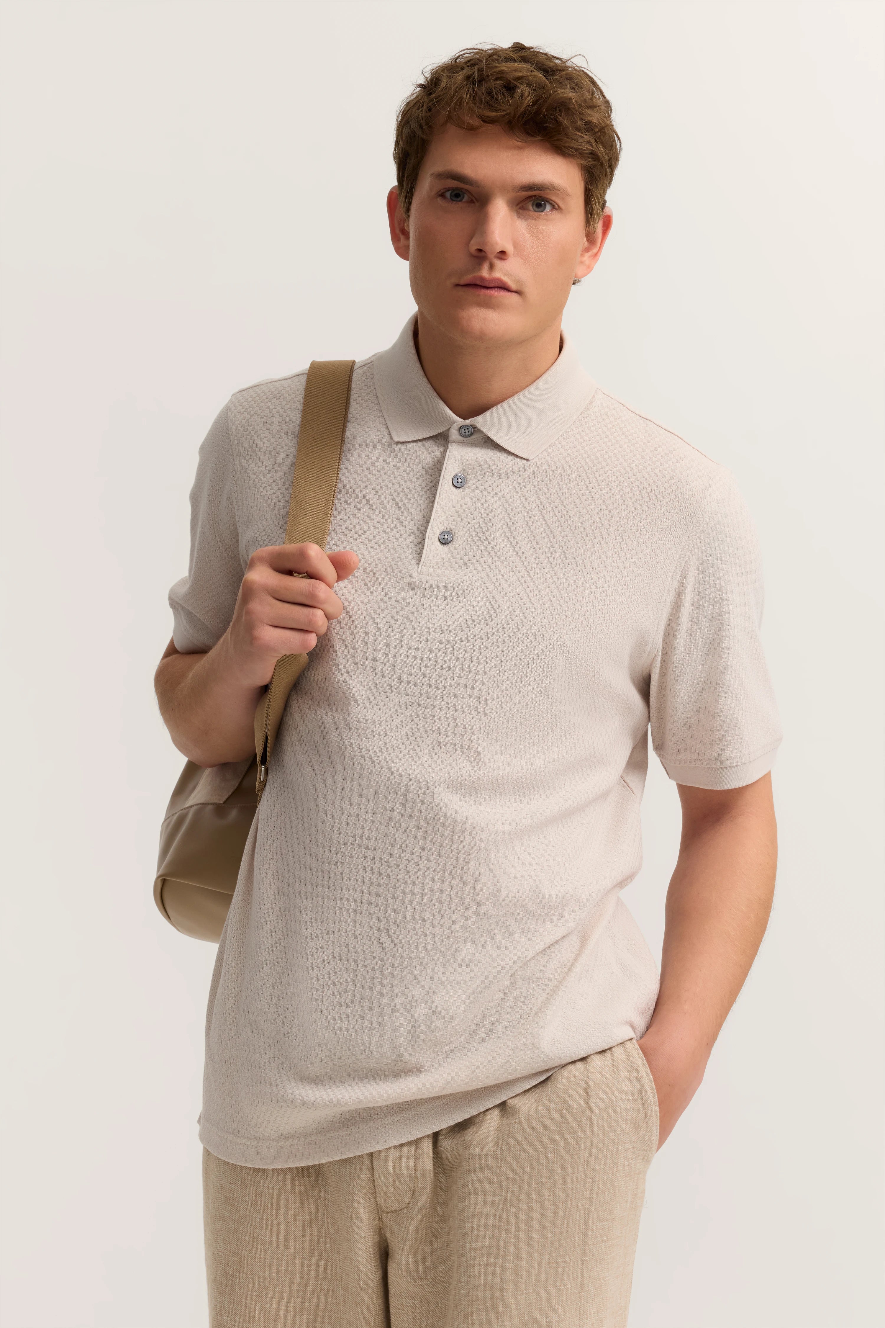 Beiges bugatti Polohemd mit Strukturmuster, beige Hose, Model frontal mit Tasche über Schulter.