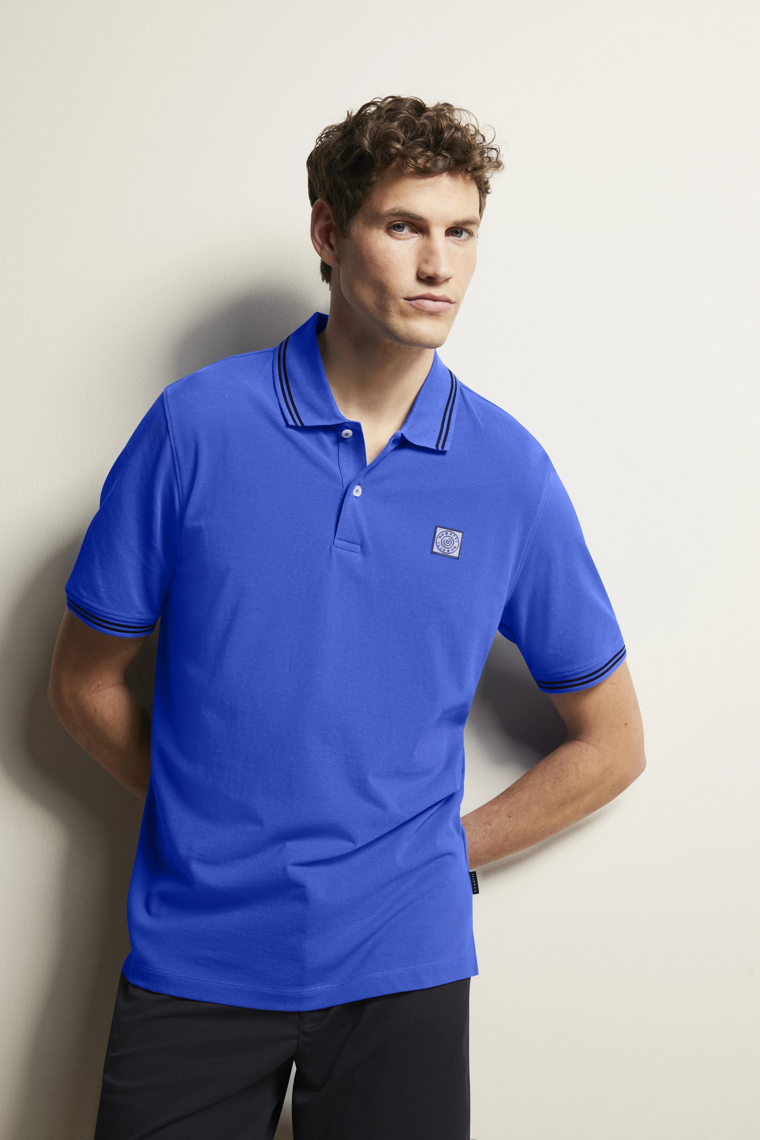 Blue bugatti polo shirt, casual model pose, relaxed vibe, black pants.