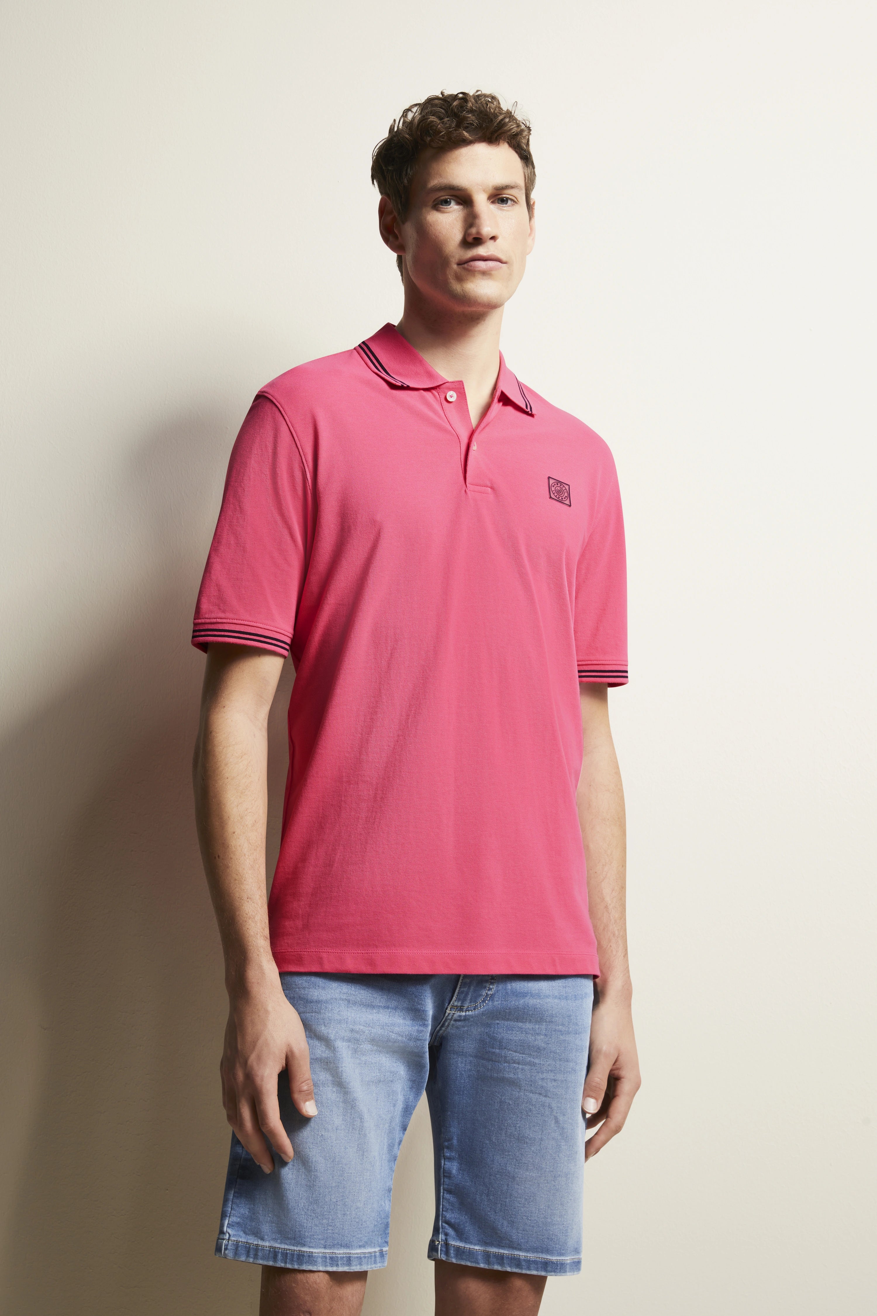 Model in pink polo shirt and blue denim shorts, casual pose, bugatti style.