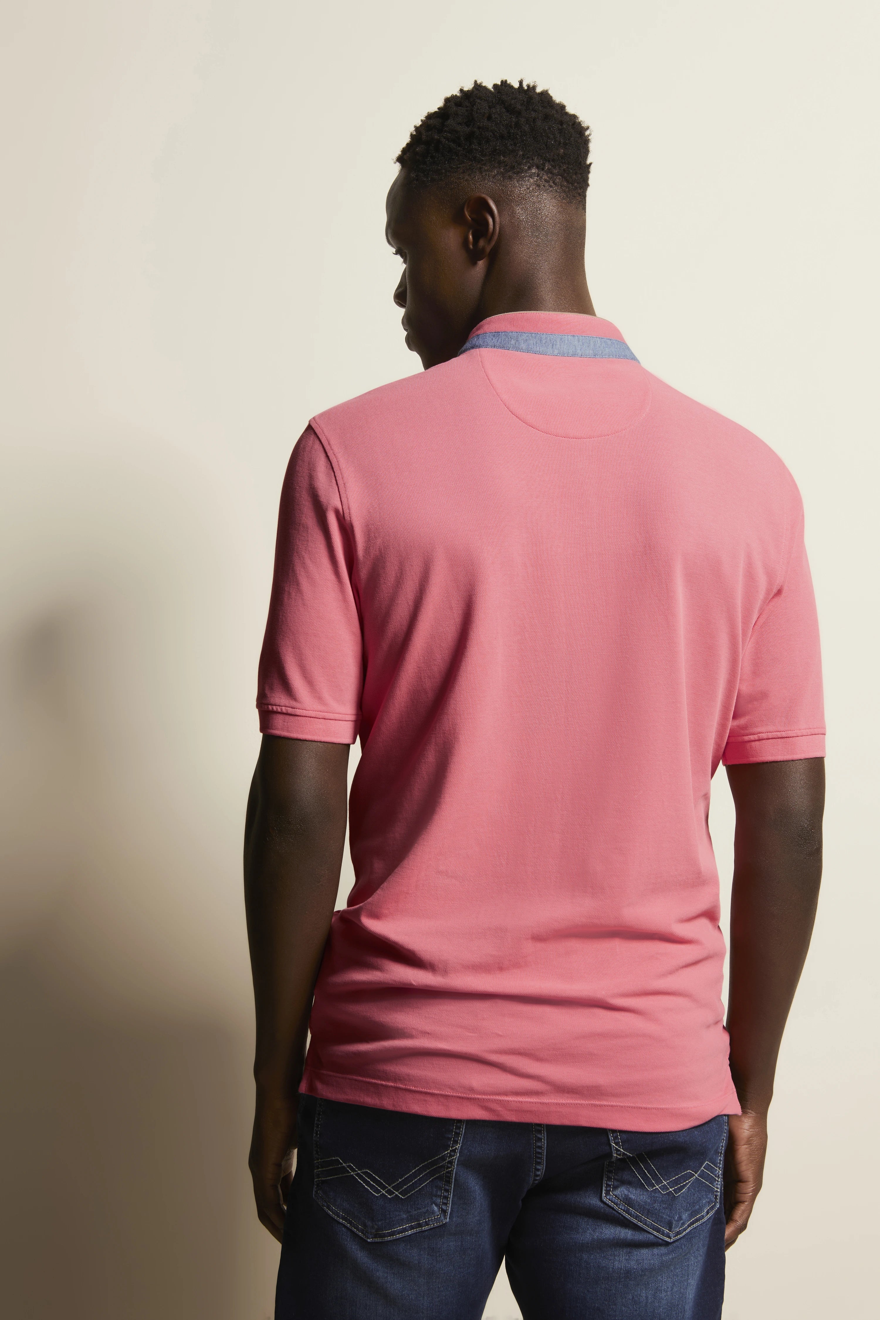 Model in bugatti roze polo en blauwe jeans van achteren, ontspannen pose.
