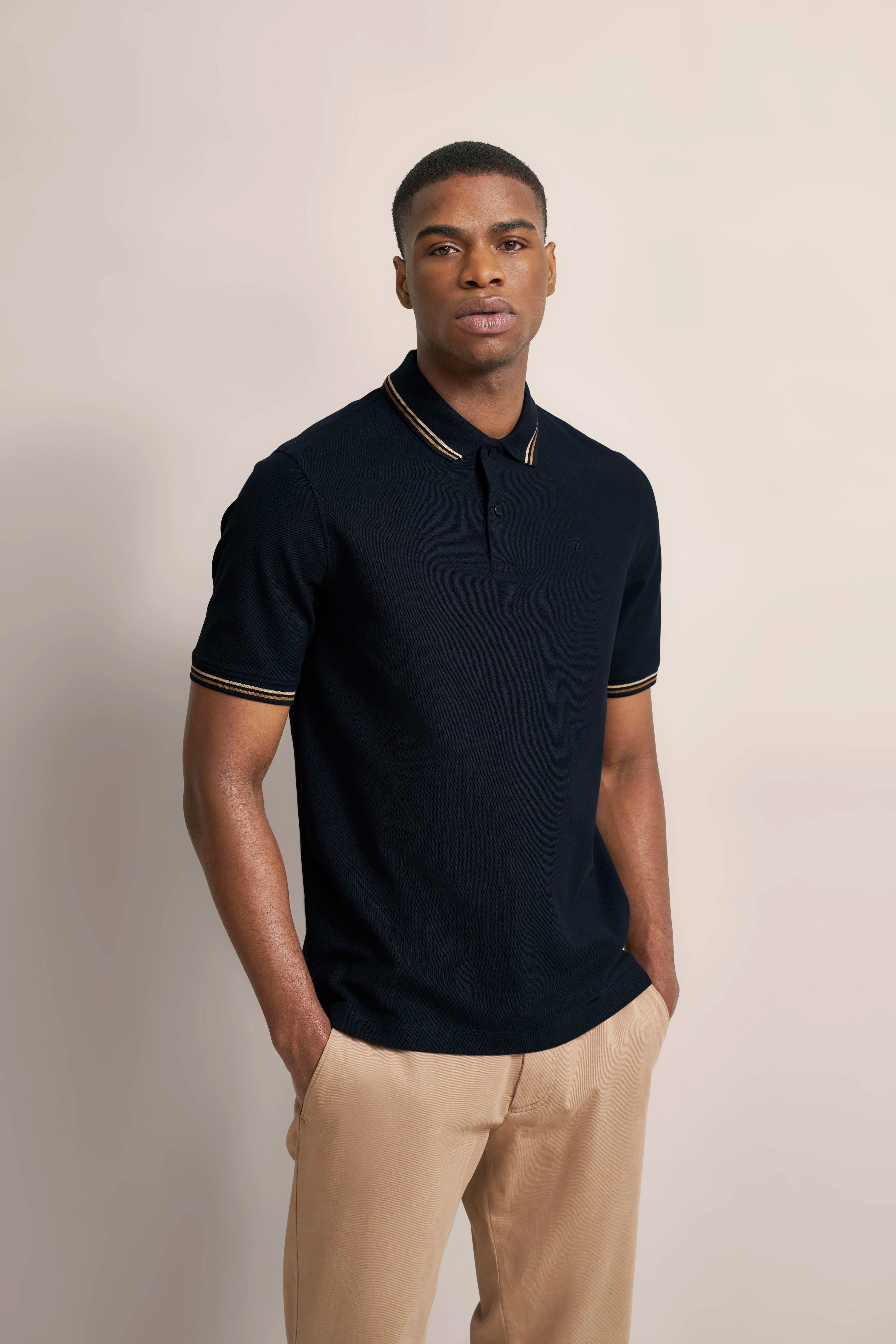 Man draagt navy poloshirt met gouden details, beige broek, ontspannen houding, geïnspireerd door Italiaanse elegantie.