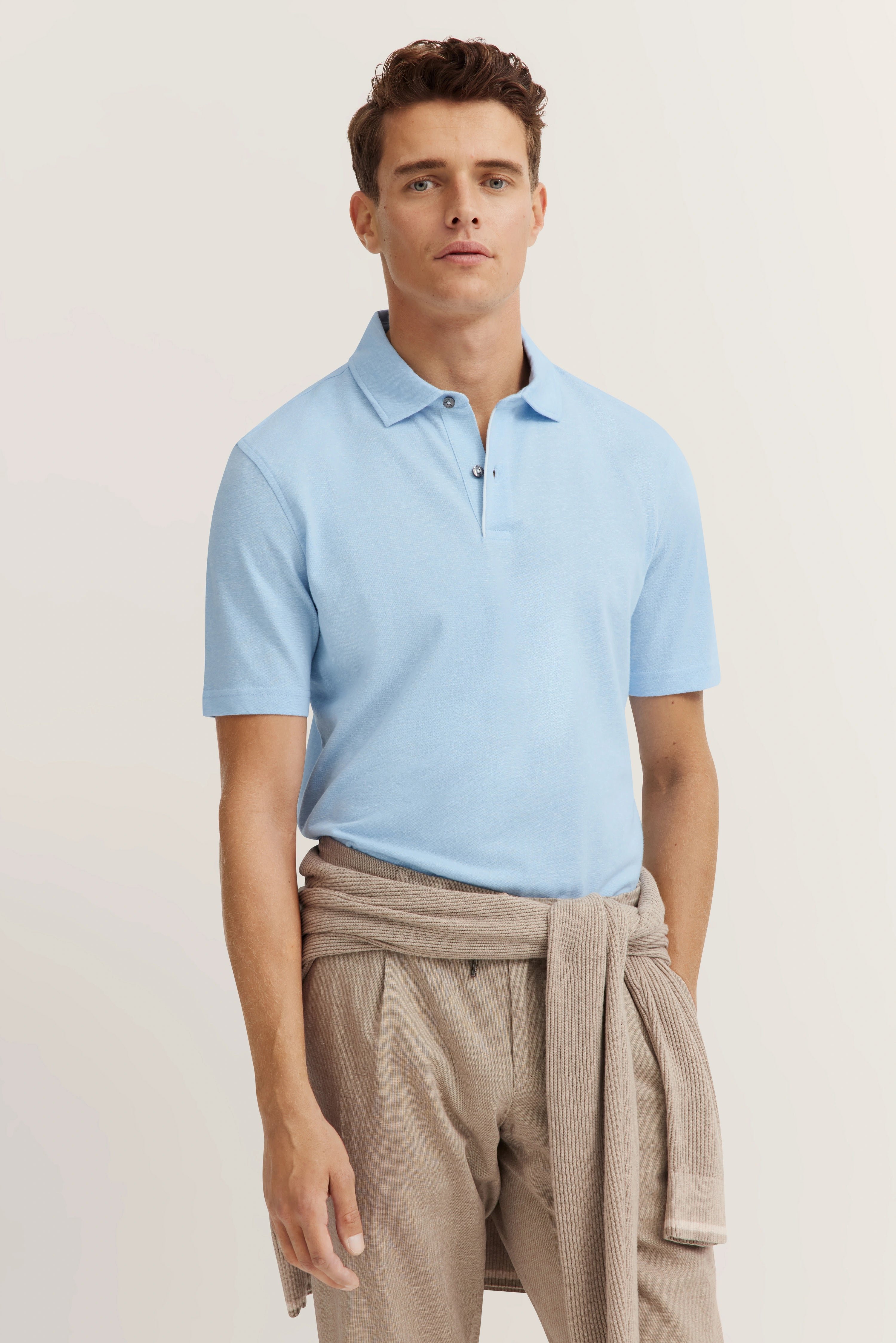 Model in light blue polo shirt, beige pants, sweater tied at waist, relaxed pose. bugatti style.