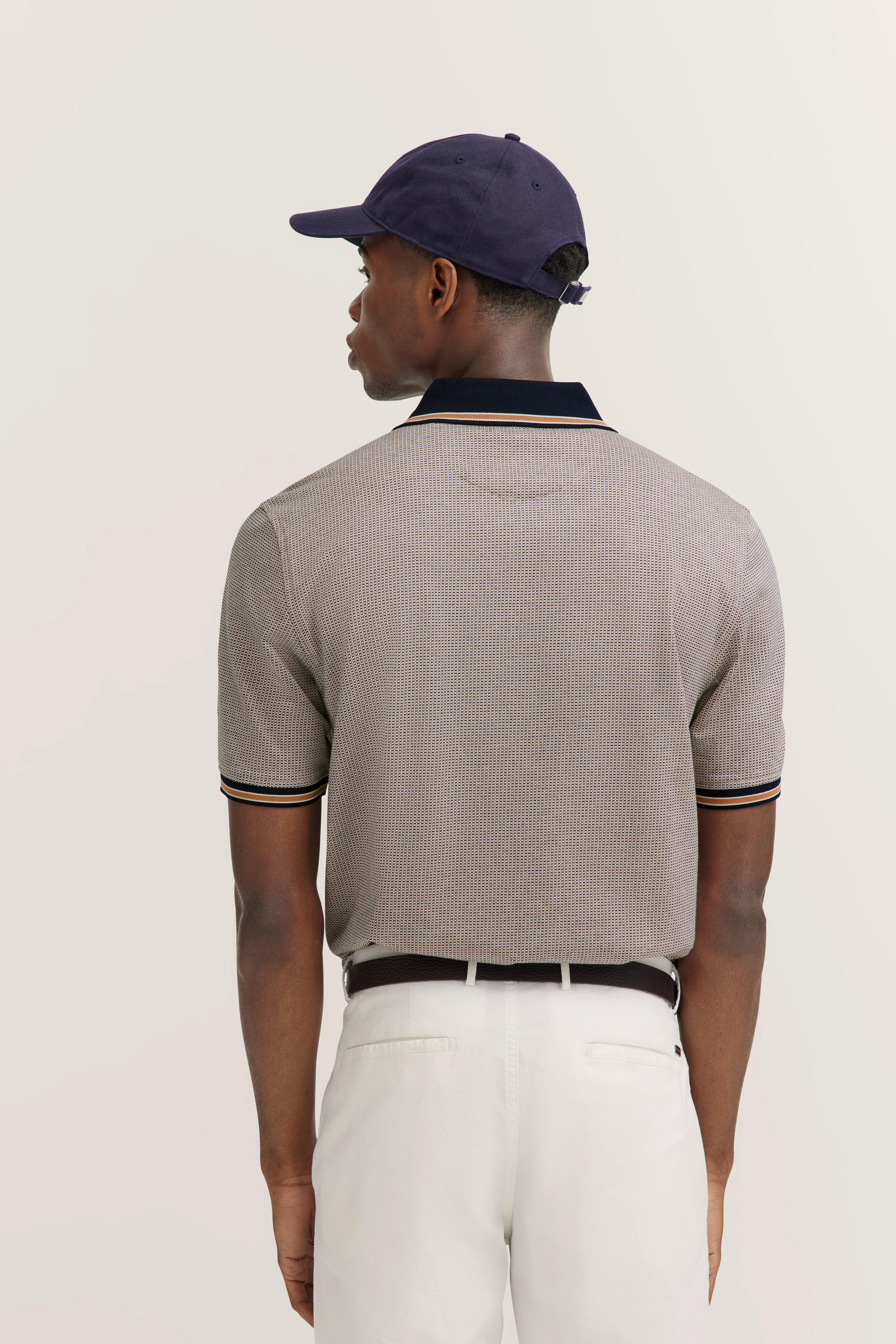 Model image, man in bugatti polo with fine pattern, beige tones, dark blue collar, white pants, relaxed pose.
