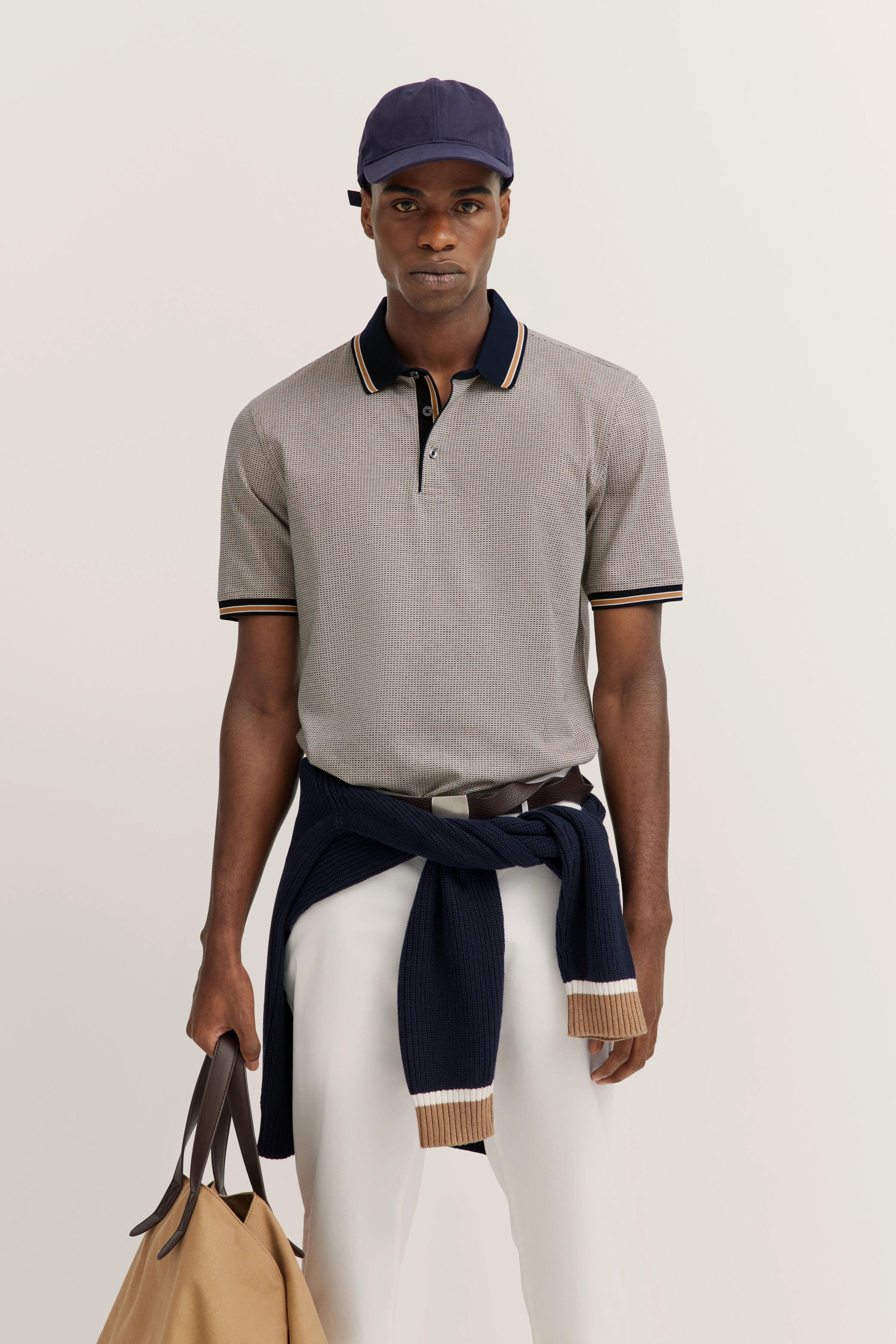 Gray polo shirt, white pants, blue sweater around waist, beige bag, model wears blue cap.