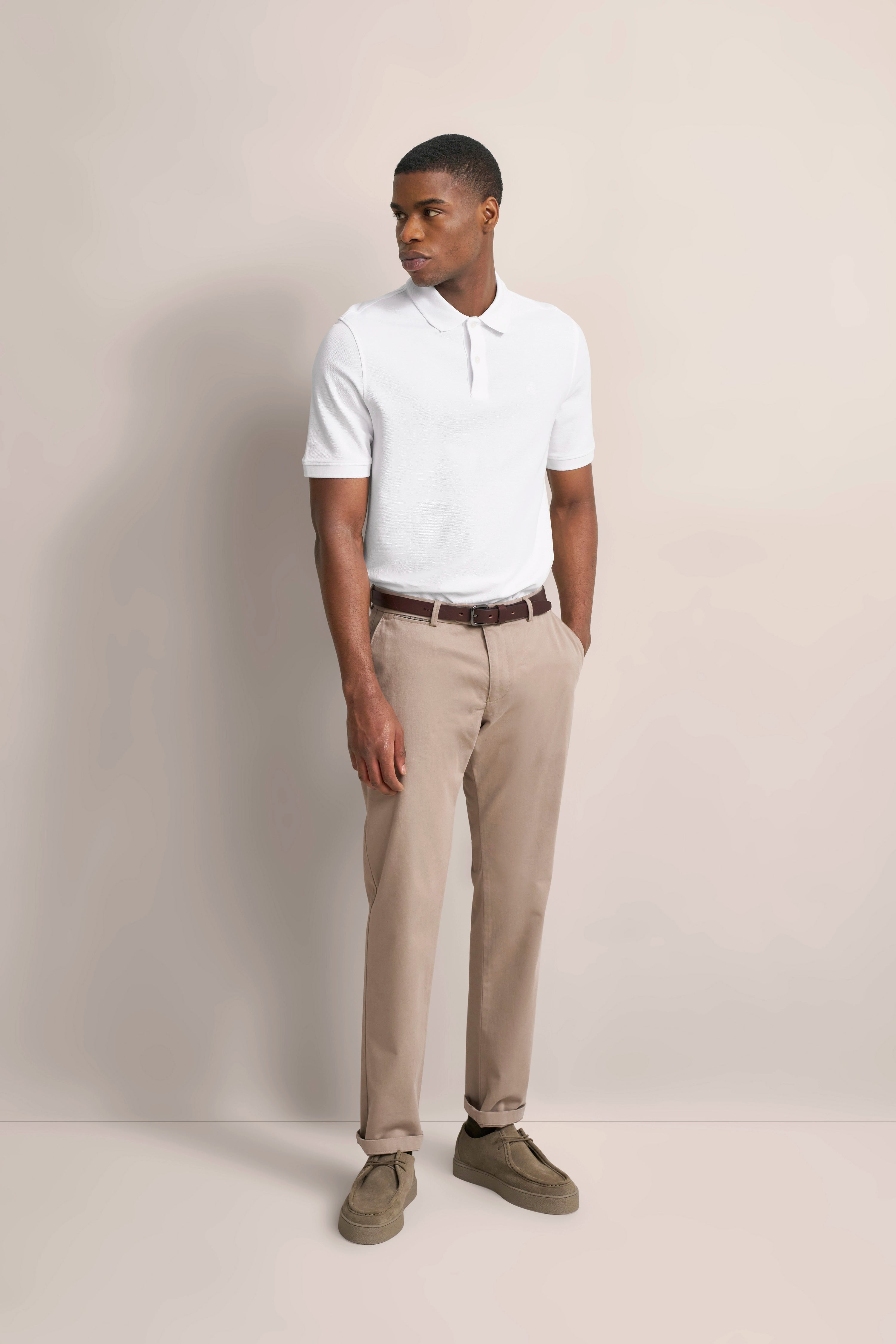Modelbild mit weißem Polo, beige Chinos und braunem Gürtel. Lässige Pose, Blick zur Seite.