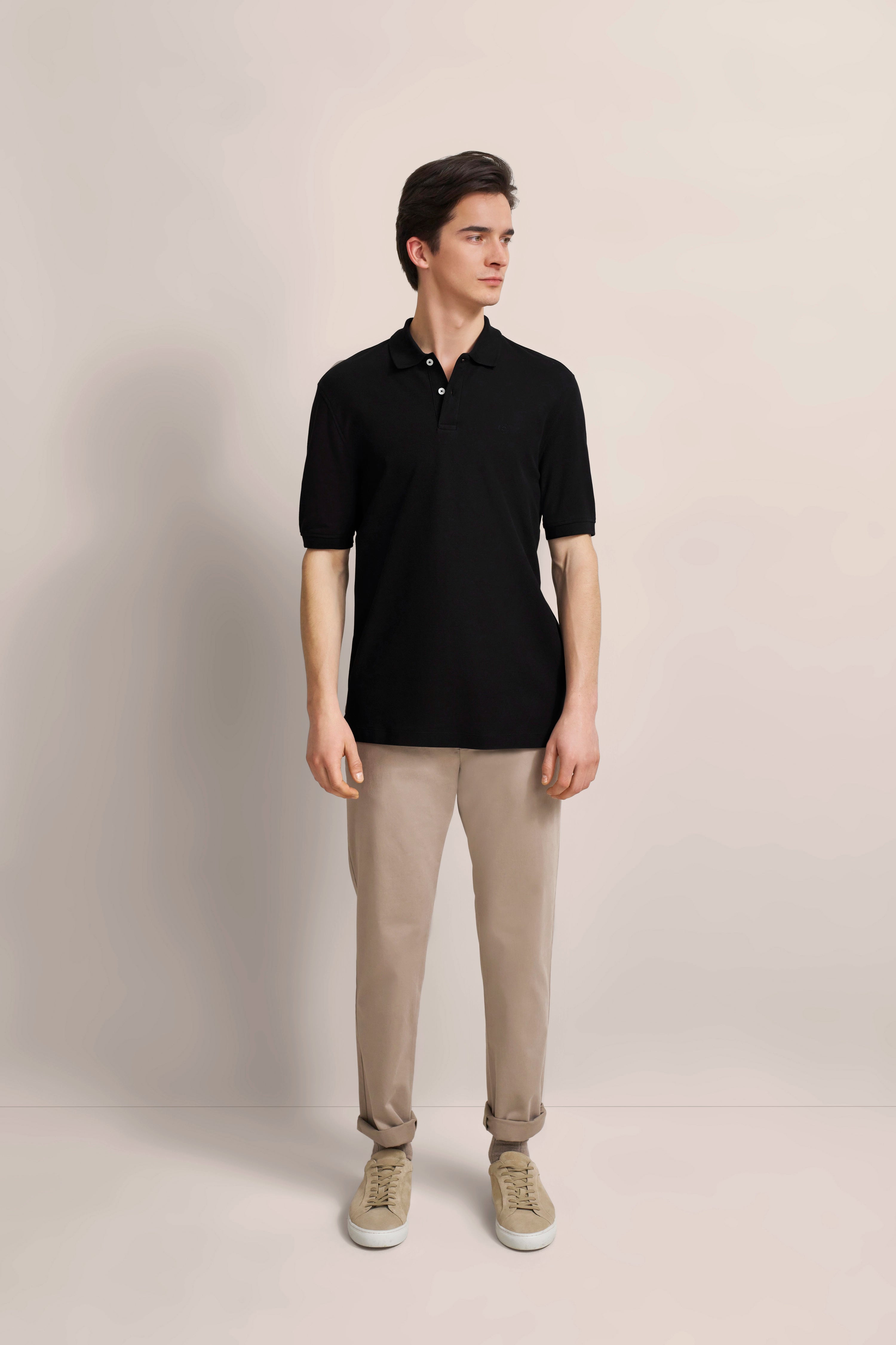 Model in black polo shirt and beige chinos, casually standing, bugatti style.