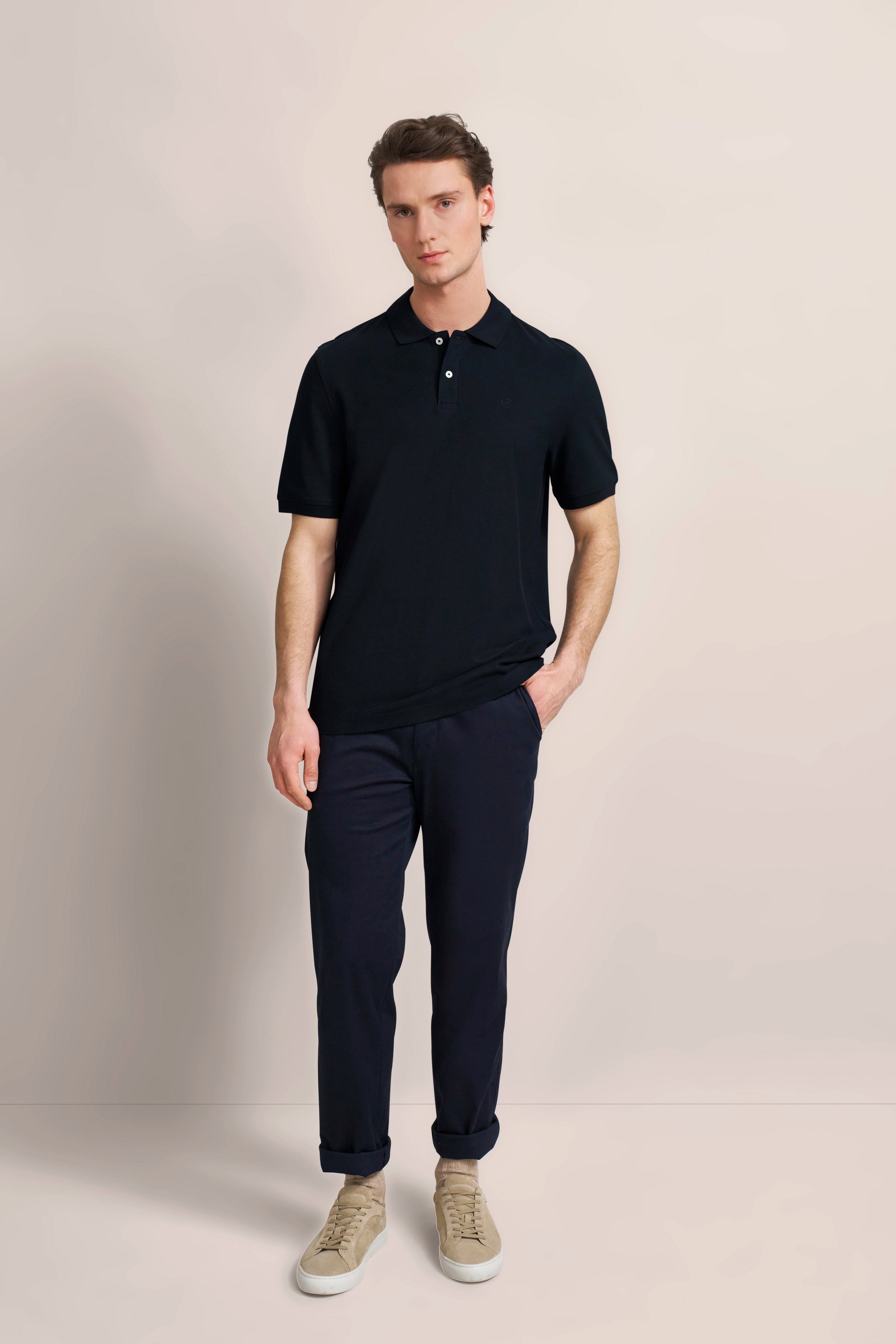 Model in zwarte polo en blauwe broek van bugatti, ontspannen pose, handen in broekzakken.