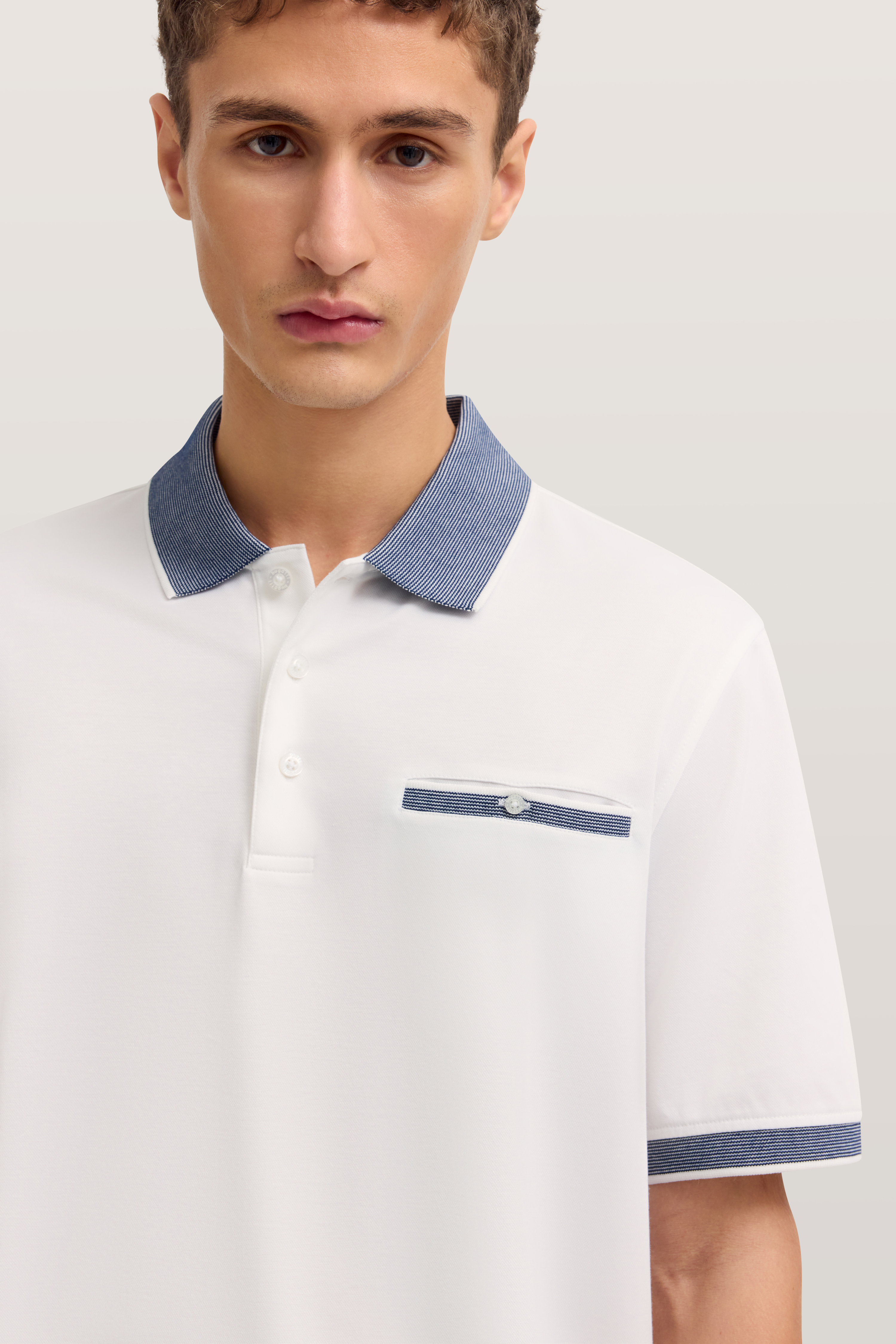 Weißes Poloshirt mit blauem Kragen und Brusttasche, Modelbild, neutraler Gesichtsausdruck.