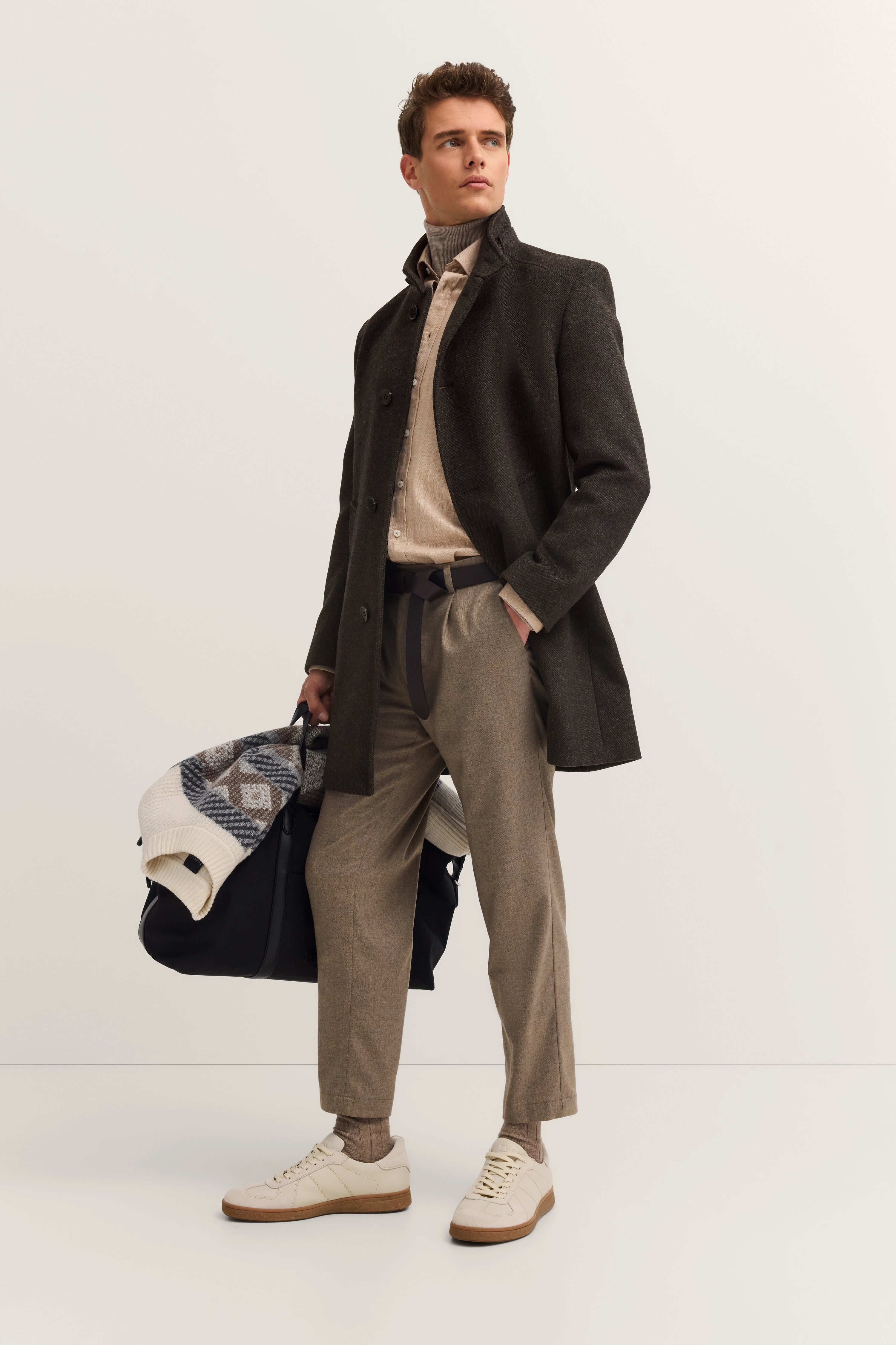 Model in grijze bugatti-jas, beige broek, overhemd. Ontspannen pose met tas.