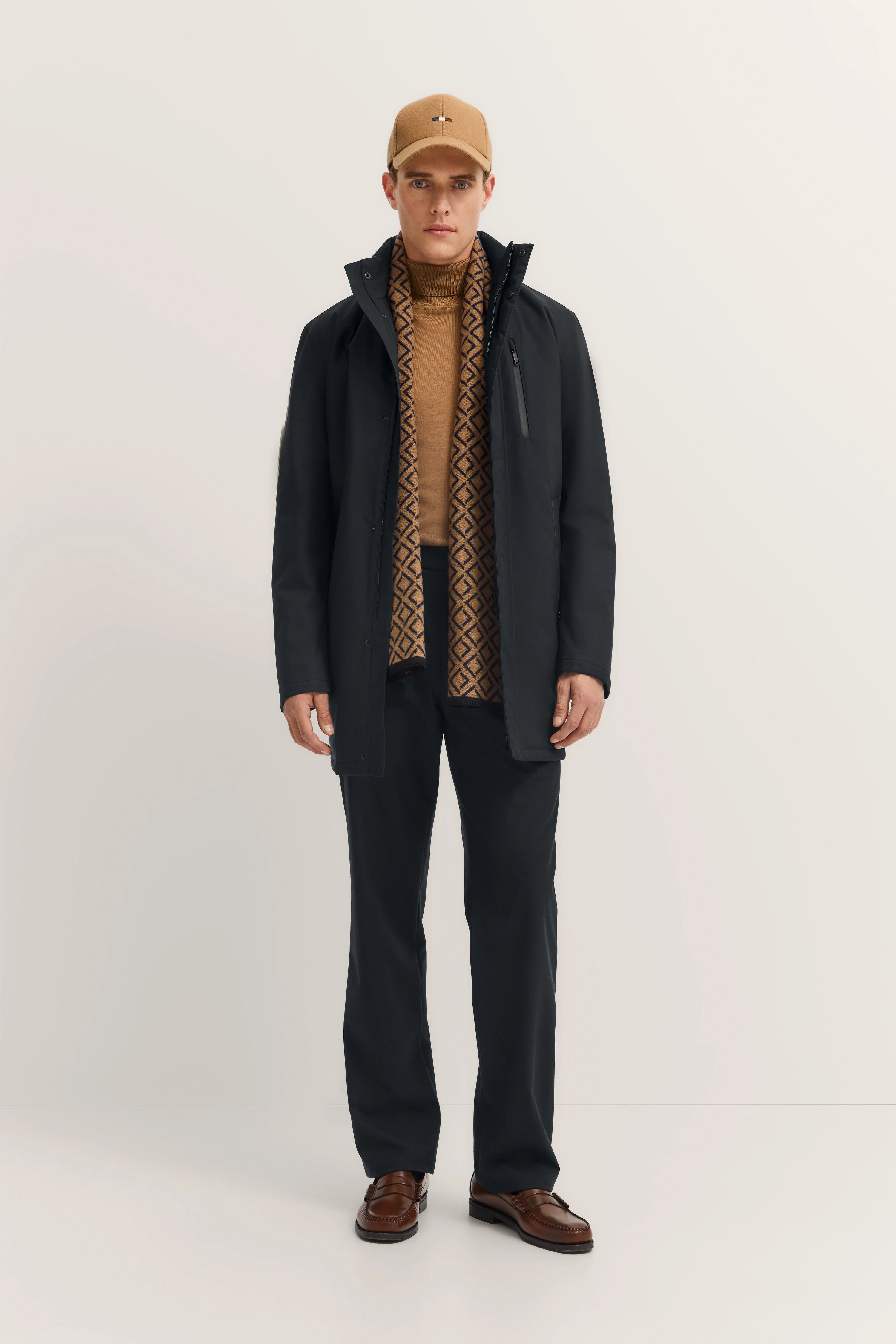 Model in schwarzem bugatti Mantel, braunem Pullover, gemustertem Schal, schwarzer Hose, braunen Schuhen, beige Cap.