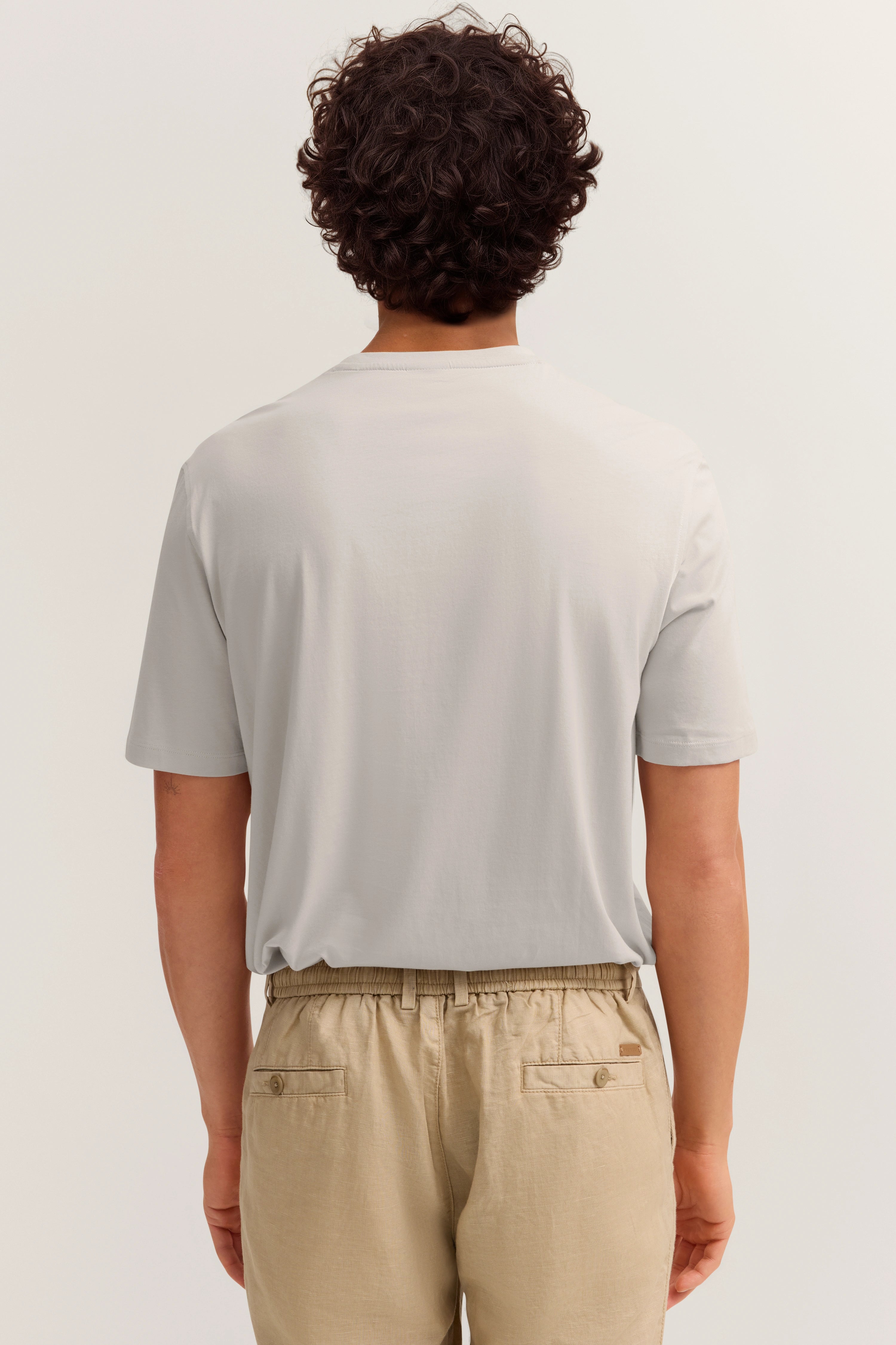 Beiges Modell in hellgrauem T-Shirt, Rückenansicht, legeres Posing.