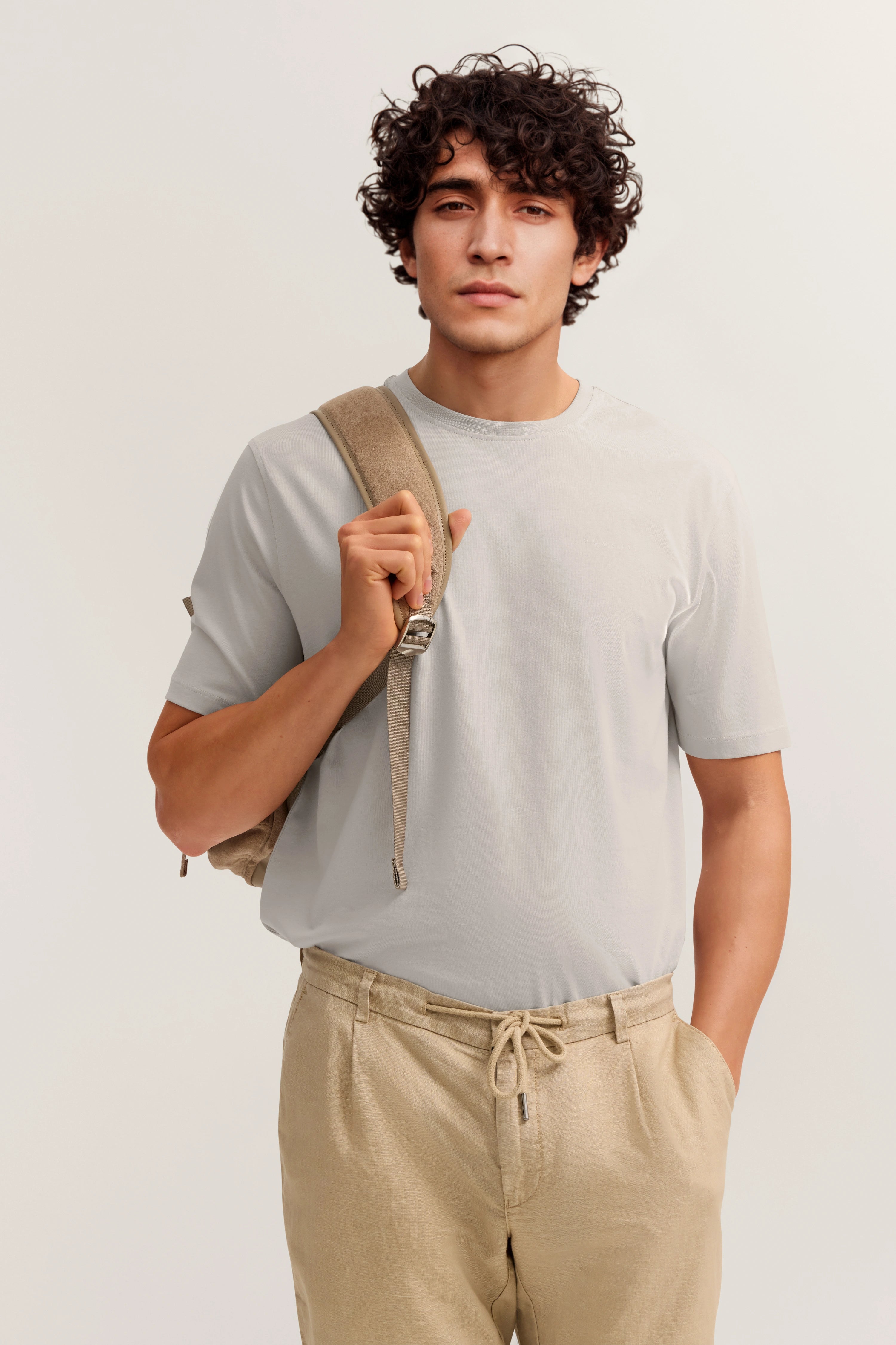 Bugatti Modelbild: Beige Hose, graues T-Shirt, Rucksack, entspannte Pose.