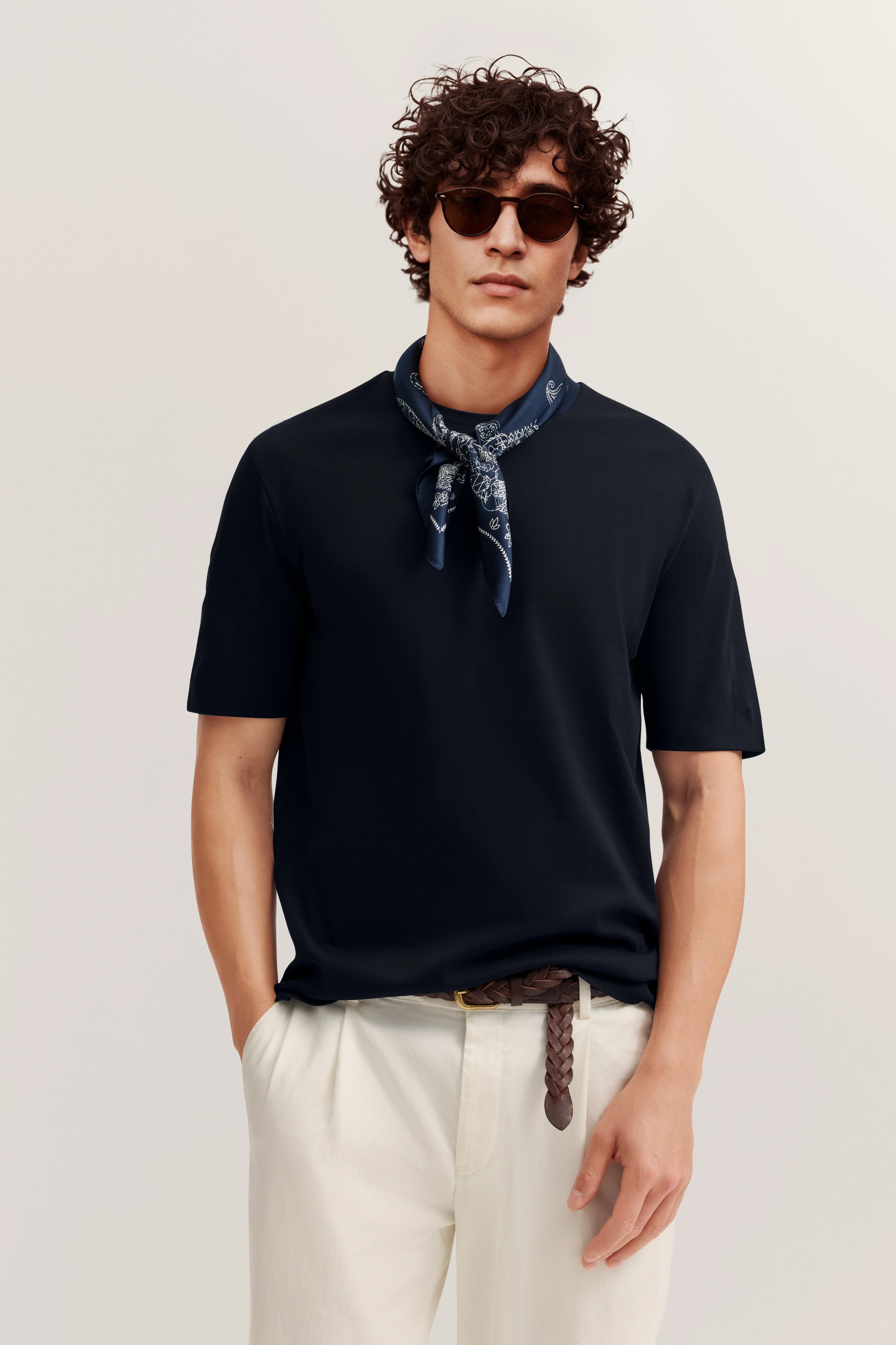 Model trägt schwarzes T-Shirt, weiße Hose, blauen Schal. Lässig, Hände in den Taschen.