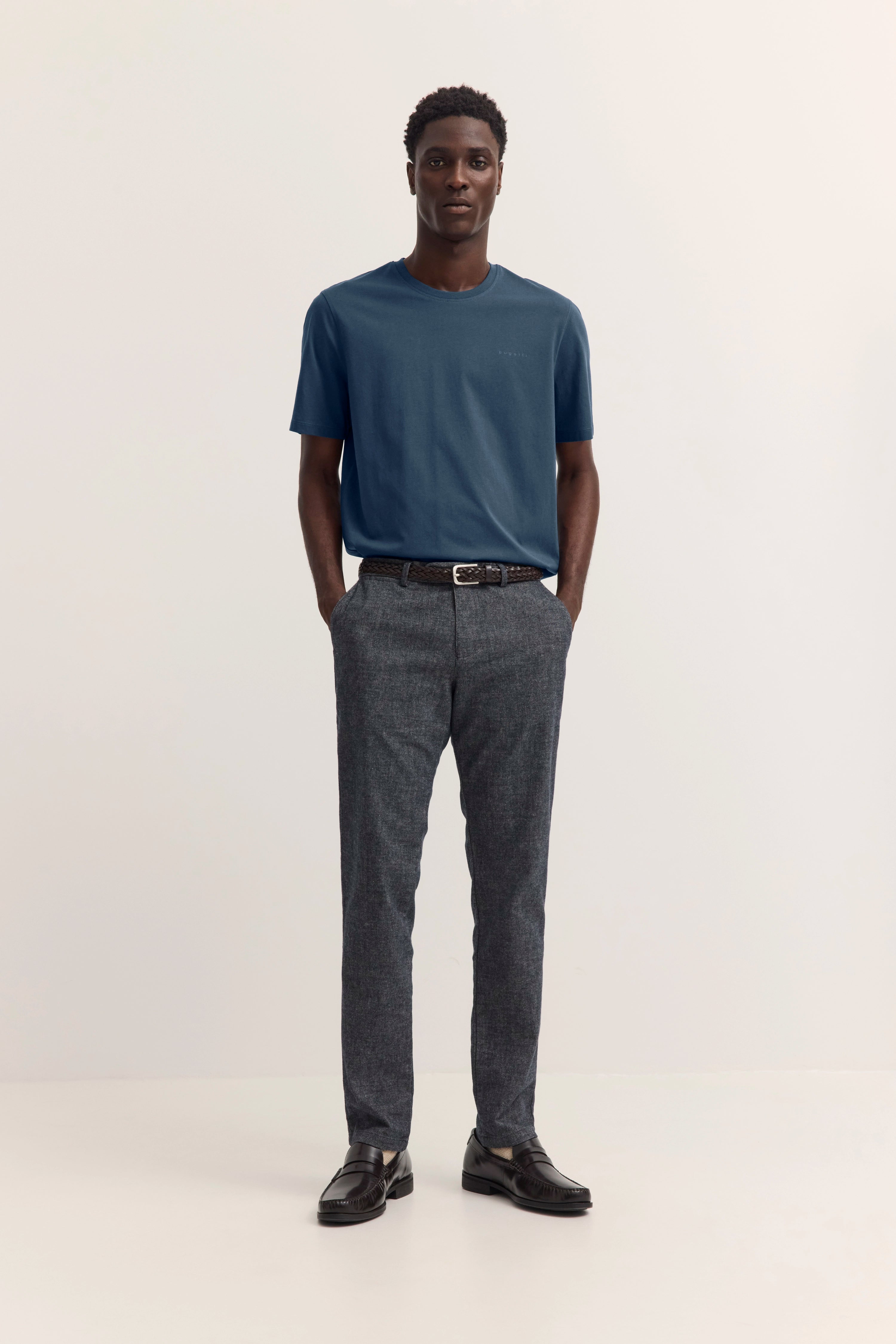 bugatti Outfit: Model in blauem T-Shirt, grauer Hose, schwarzem Gürtel, schwarze Schuhe, legeres Posing.
