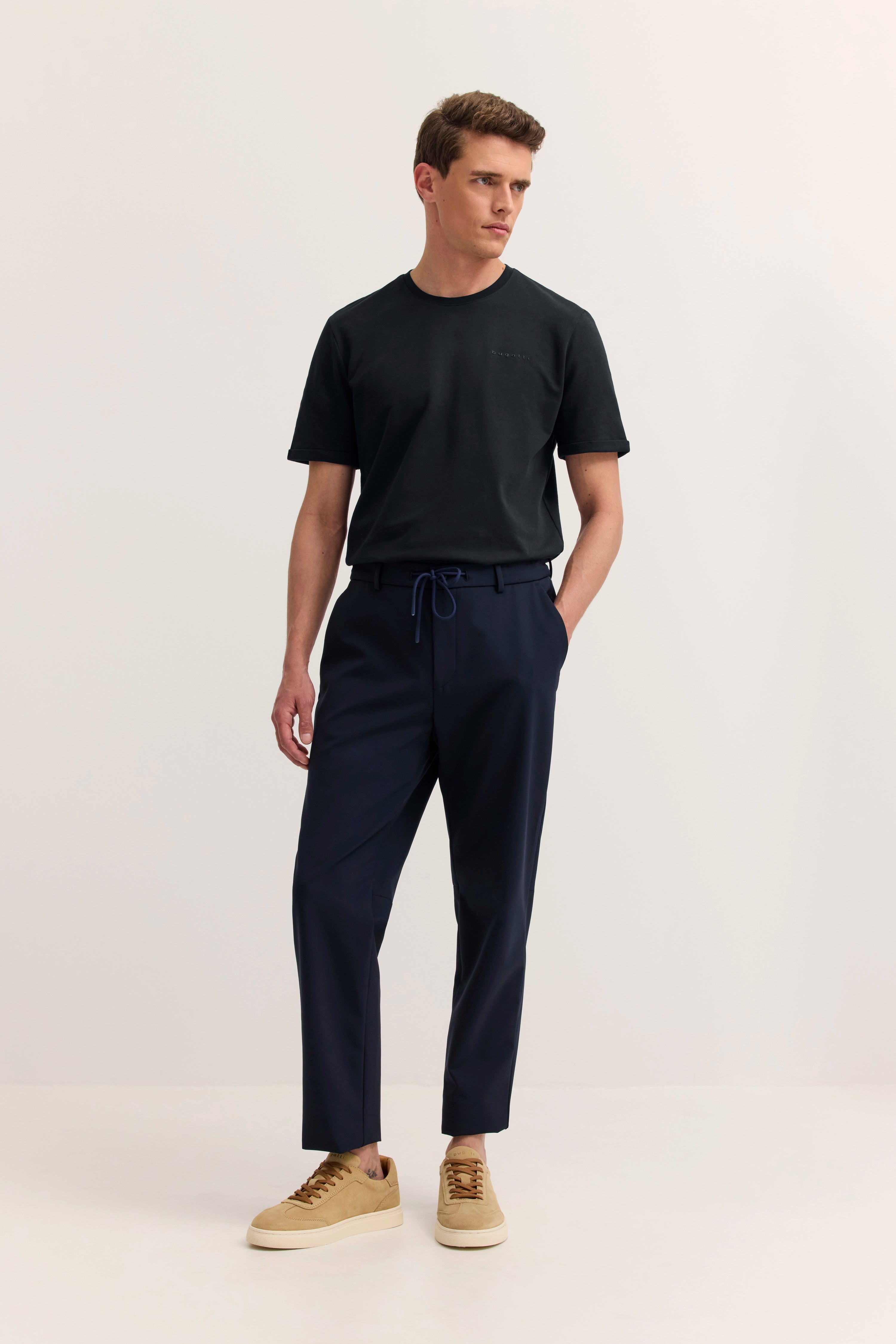 Model in schwarzem bugatti T-Shirt, blauer Hose, beigem Sneaker, entspanntes Posing.