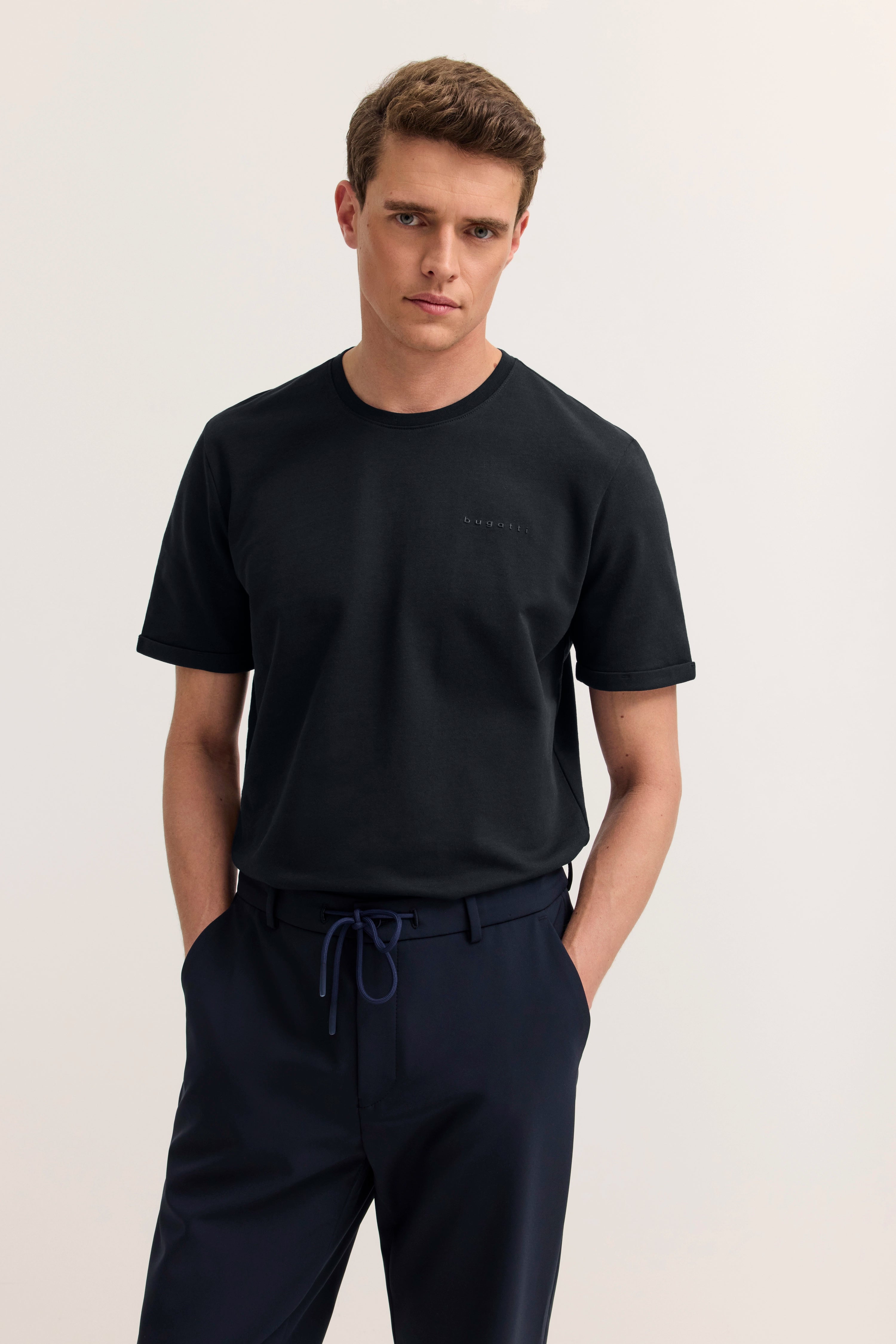 Model in schwarzem bugatti T-Shirt und marineblauer Hose, lässig stehend, Hände in den Taschen.
