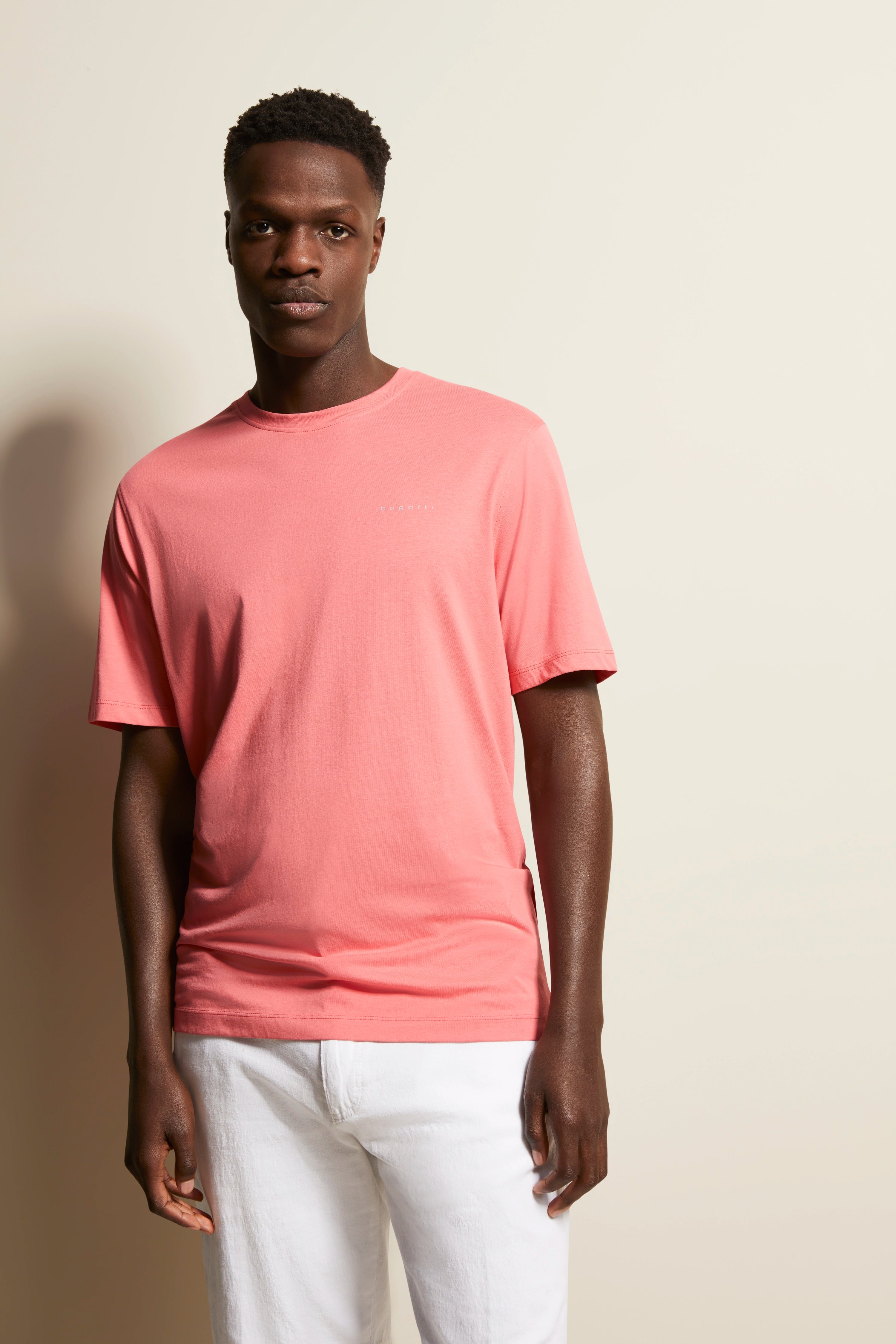 bugatti Modelbild, rosa T-Shirt, weiße Hose, entspannte Pose, zeitlose Eleganz und Stil.