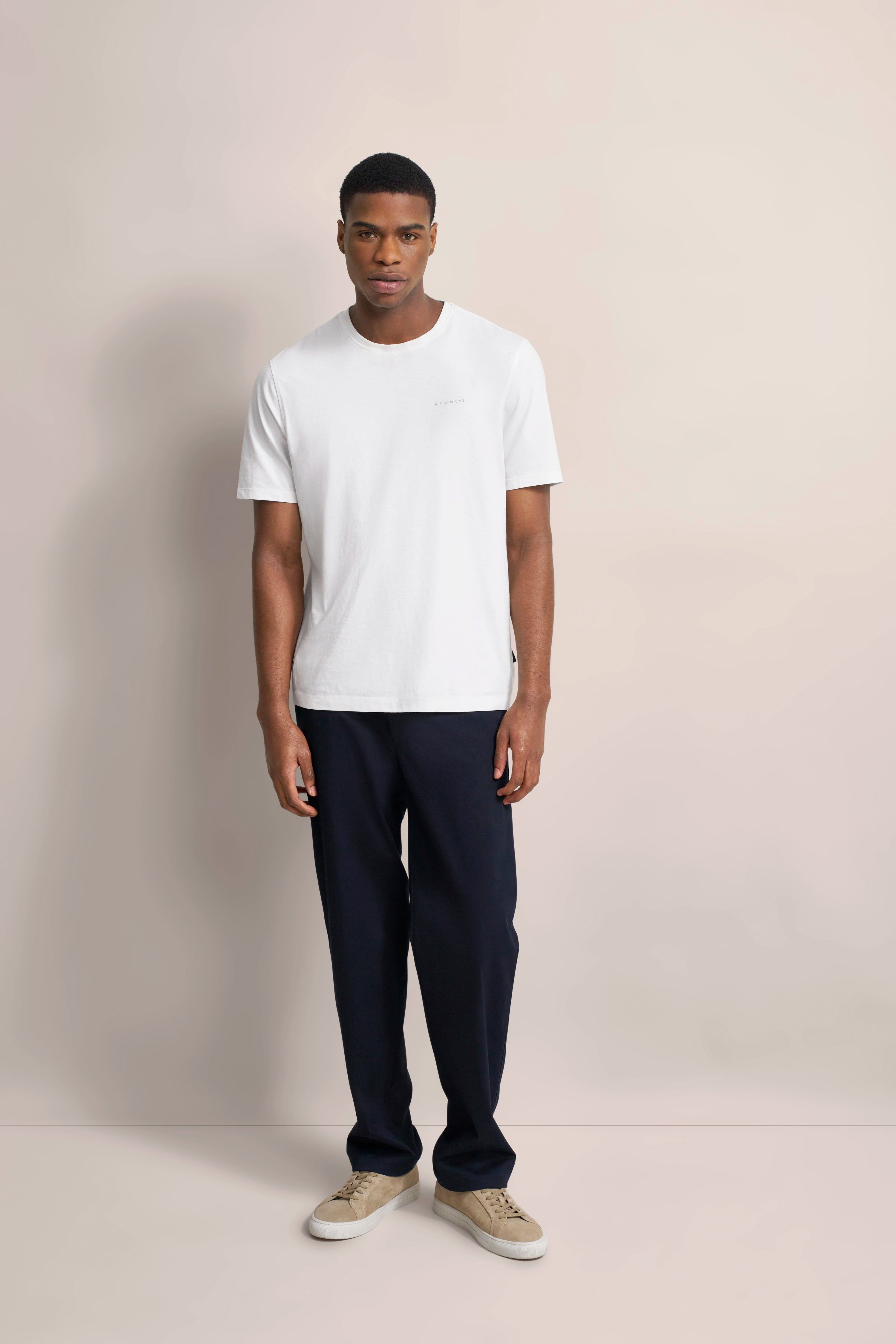 Weißes T-Shirt von bugatti, dunkelblaue Hose, beige Sneaker. Model in entspannter Pose.
