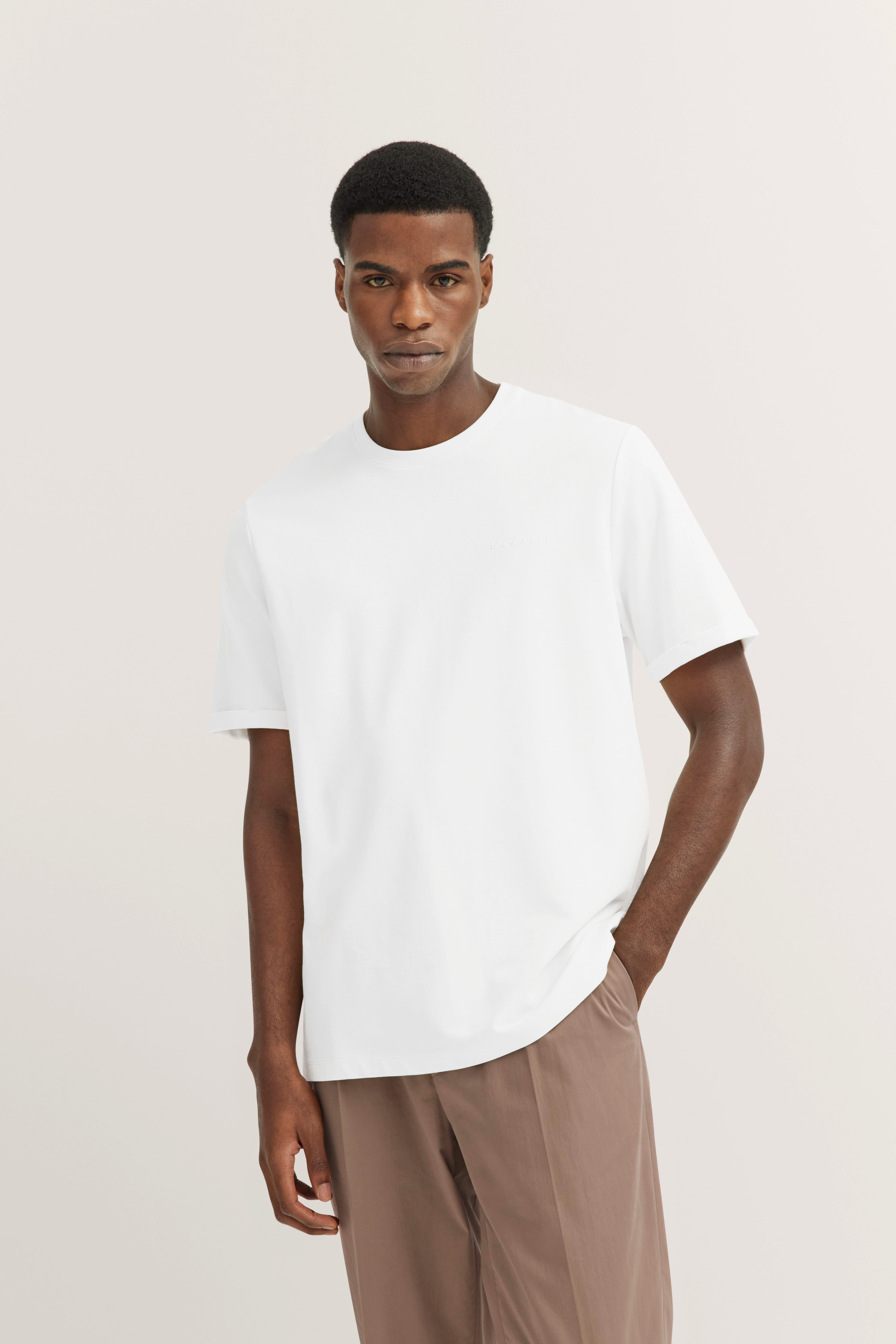 White bugatti T-shirt, brown pants, casual model image, relaxed pose.