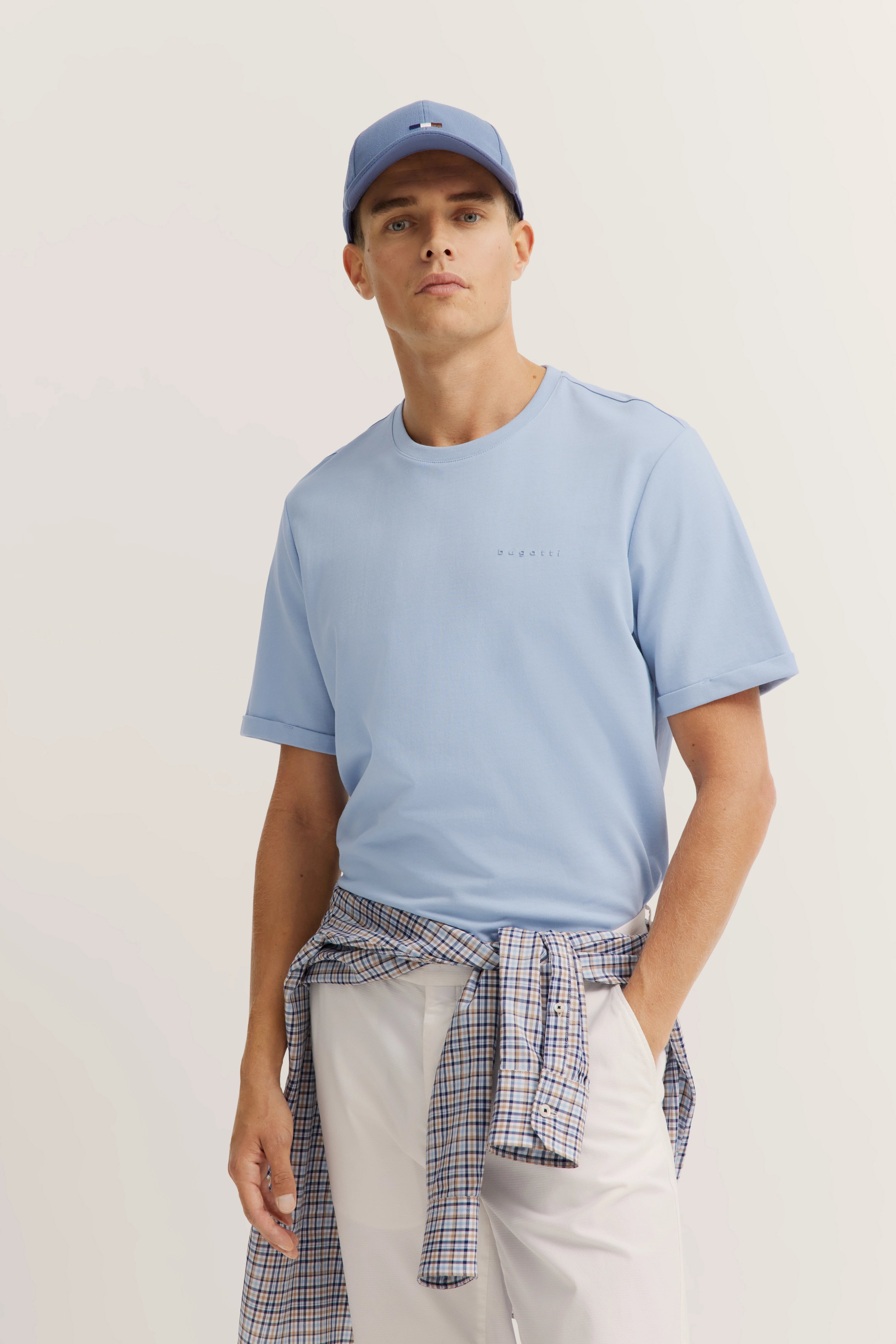 Model in light blue bugatti T-shirt, matching cap, white pants, plaid shirt tied at waist.