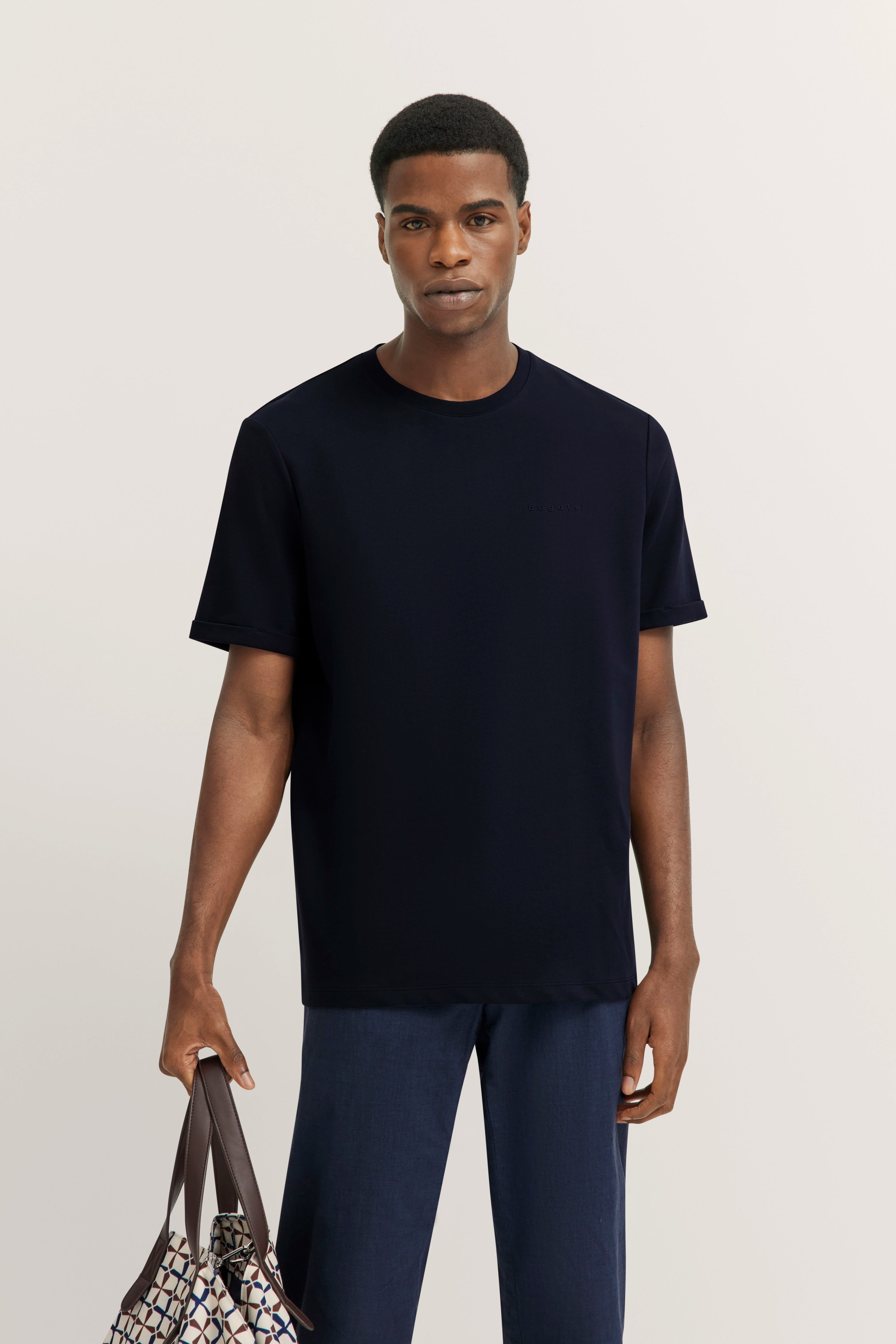 bugatti model in navy T-shirt, dark blue pants, standing, holding patterned bag.