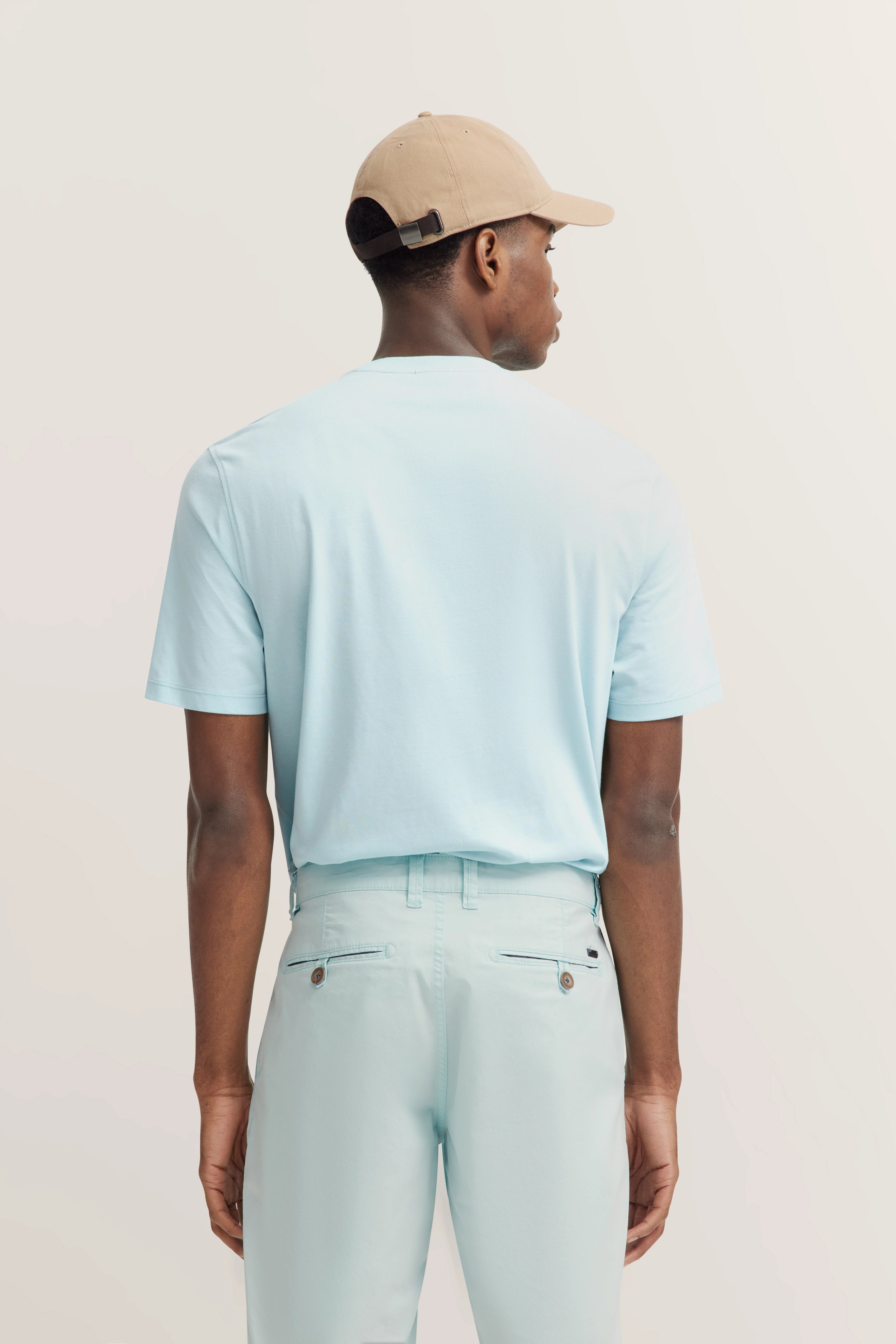 Rear view of model in light blue bugatti T-shirt and pants, wearing beige cap.