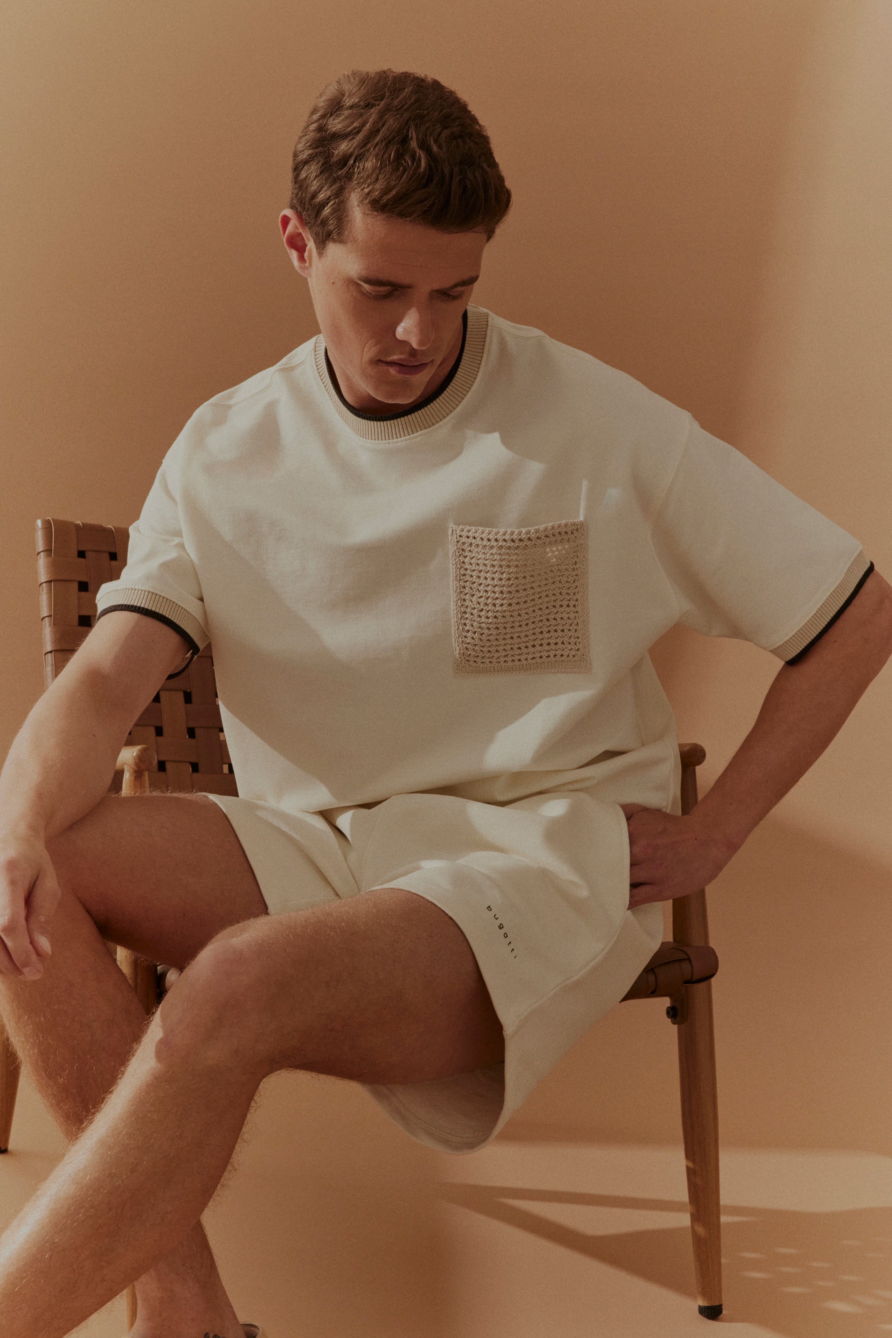 Entspannter Look mit cremefarbenem Shirt und Shorts von bugatti, Model sitzt lässig auf Stuhl.