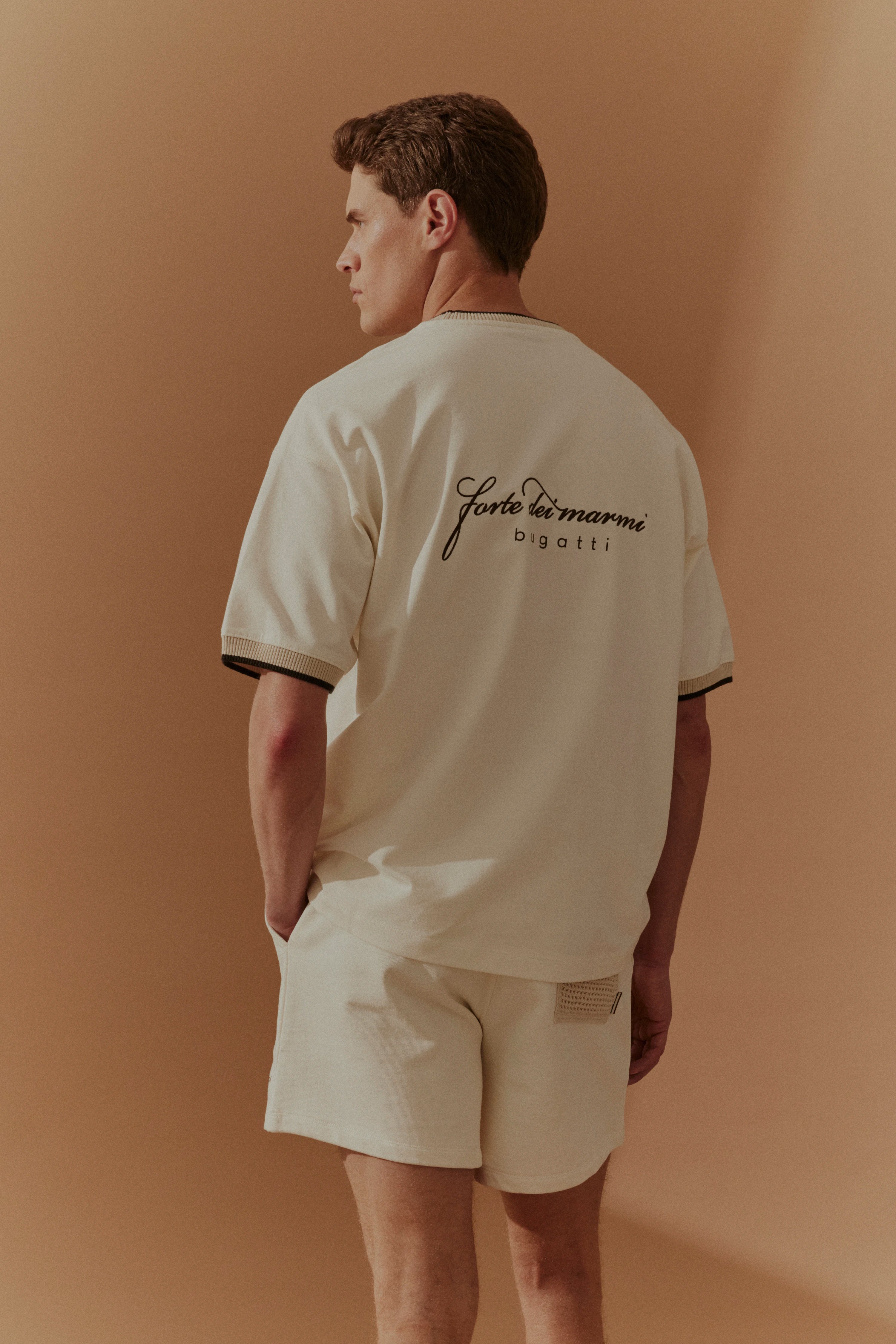 Model in cremefarbenem bugatti Outfit, lässige Pose, T-Shirt und Shorts mit Schriftzug hinten.