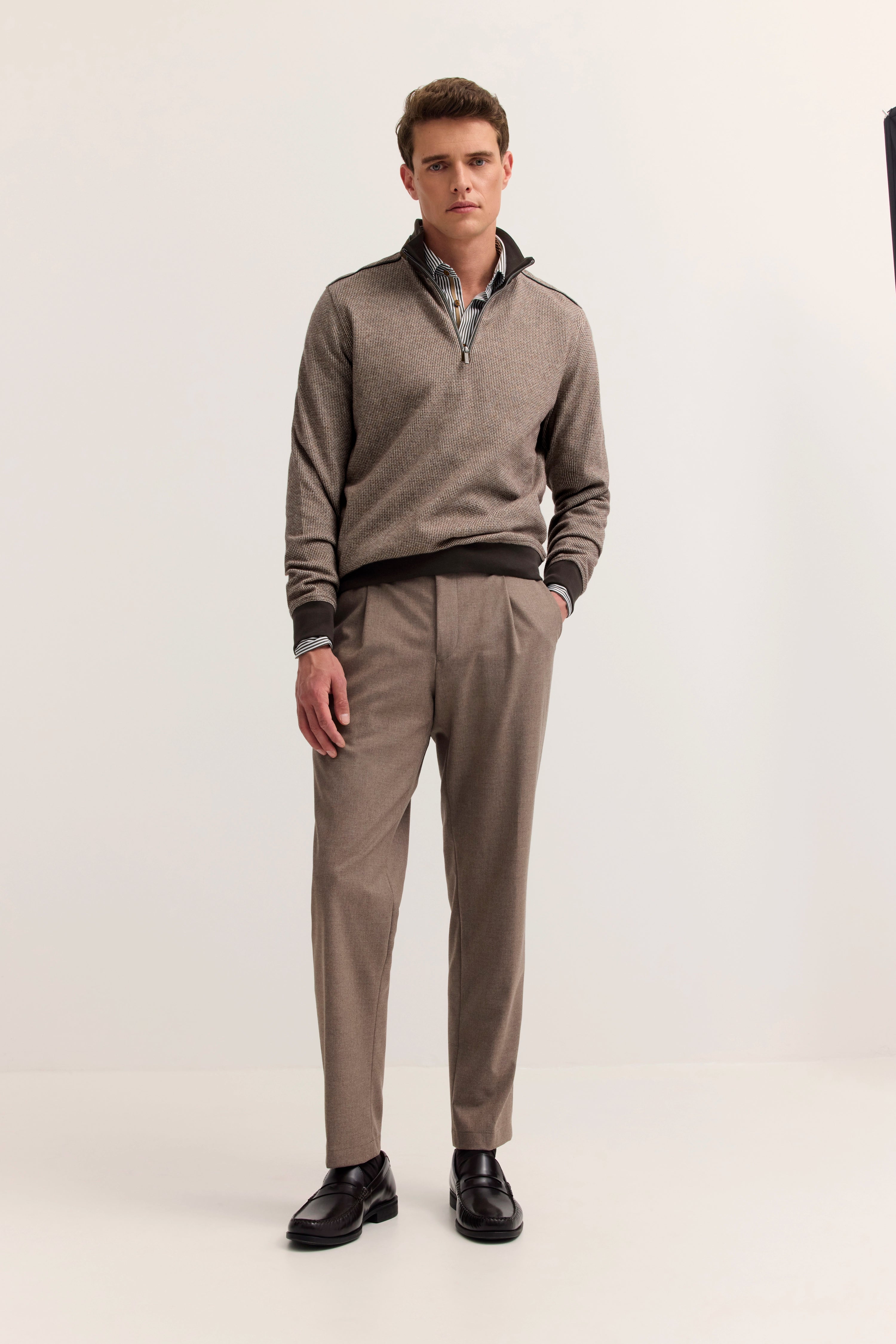 Model in bugatti-Outfit: beige Pullover, braune Hose, schwarze Schuhe, entspannte Haltung.