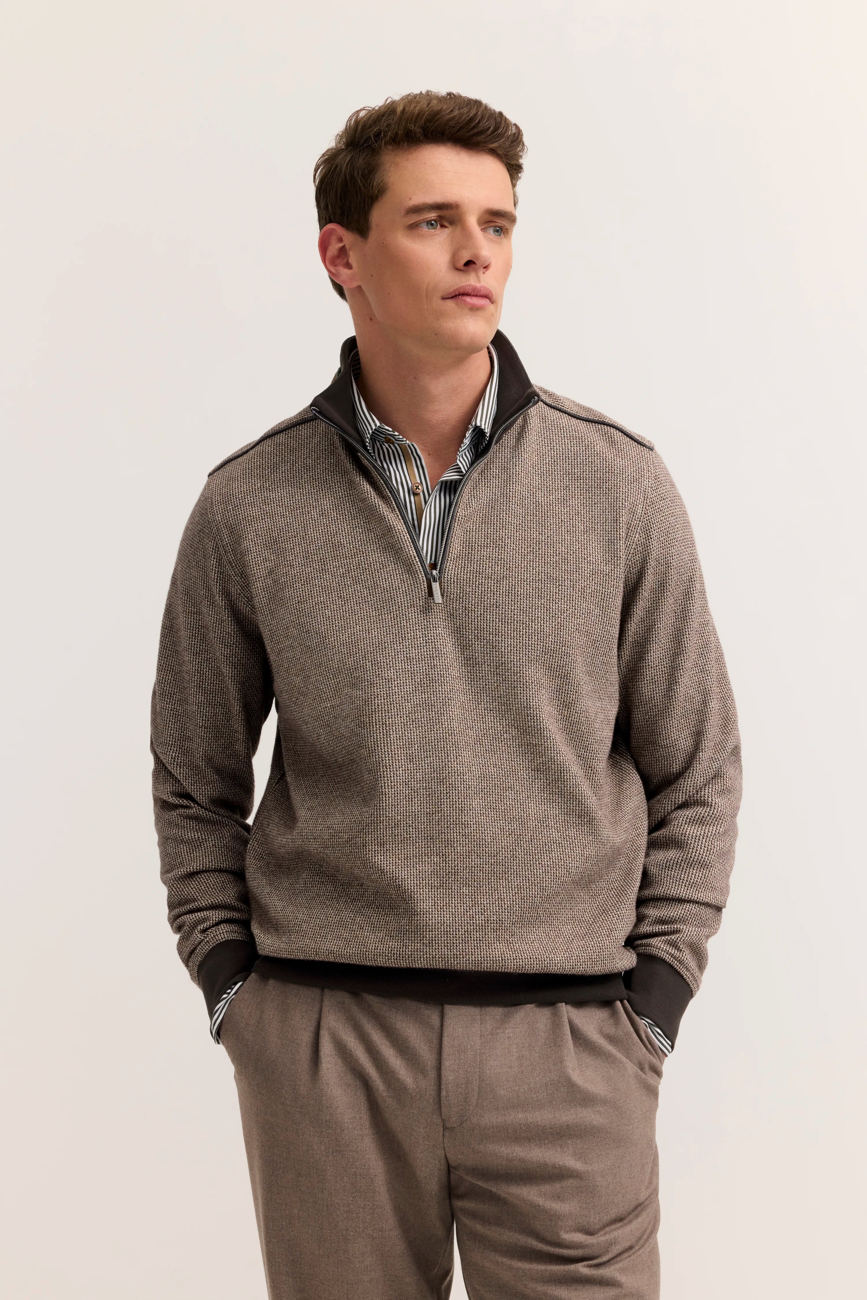 Model in beigefarbenem bugatti Pullover mit Reißverschluss, gestreiftem Hemd, brauner Hose, entspannte Pose.