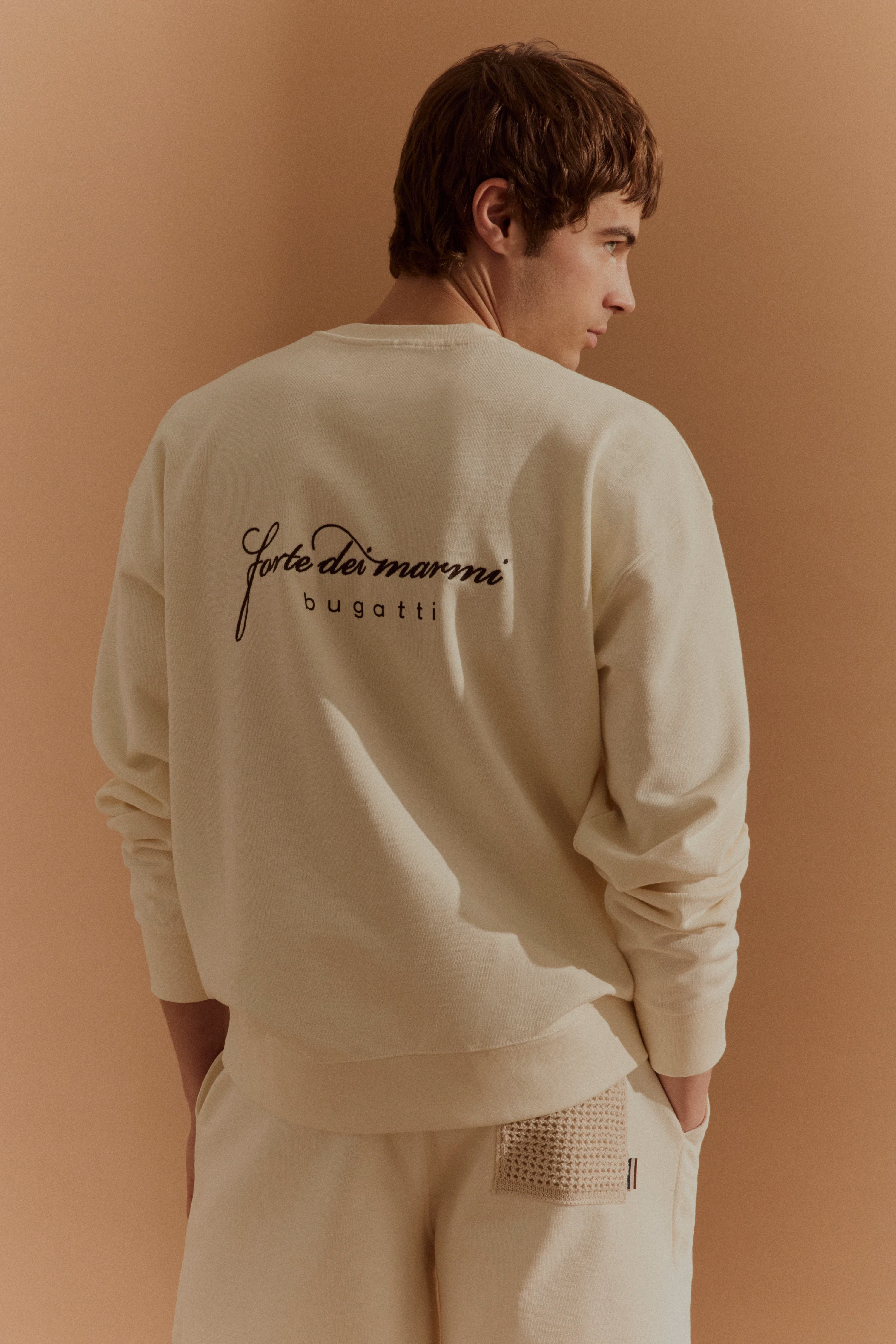 bugatti Modelbild: Cremefarbenes Sweatshirt mit Schriftzug, lässig seitlich stehend, entspannter Stil.
