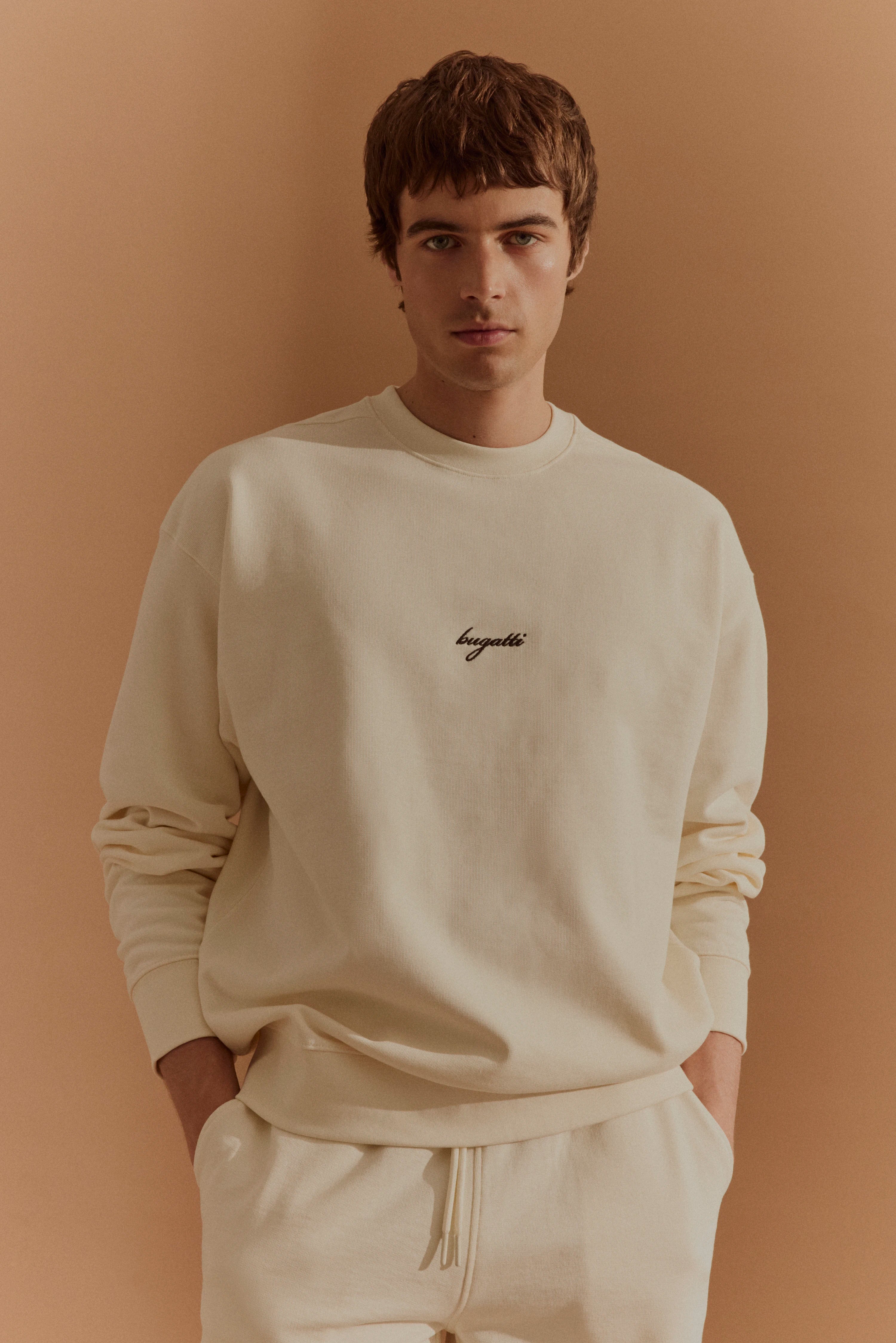 Model in cremefarbenem bugatti Sweatshirt und Hose vor beige Hintergrund, lässige Pose, Hände in Taschen.