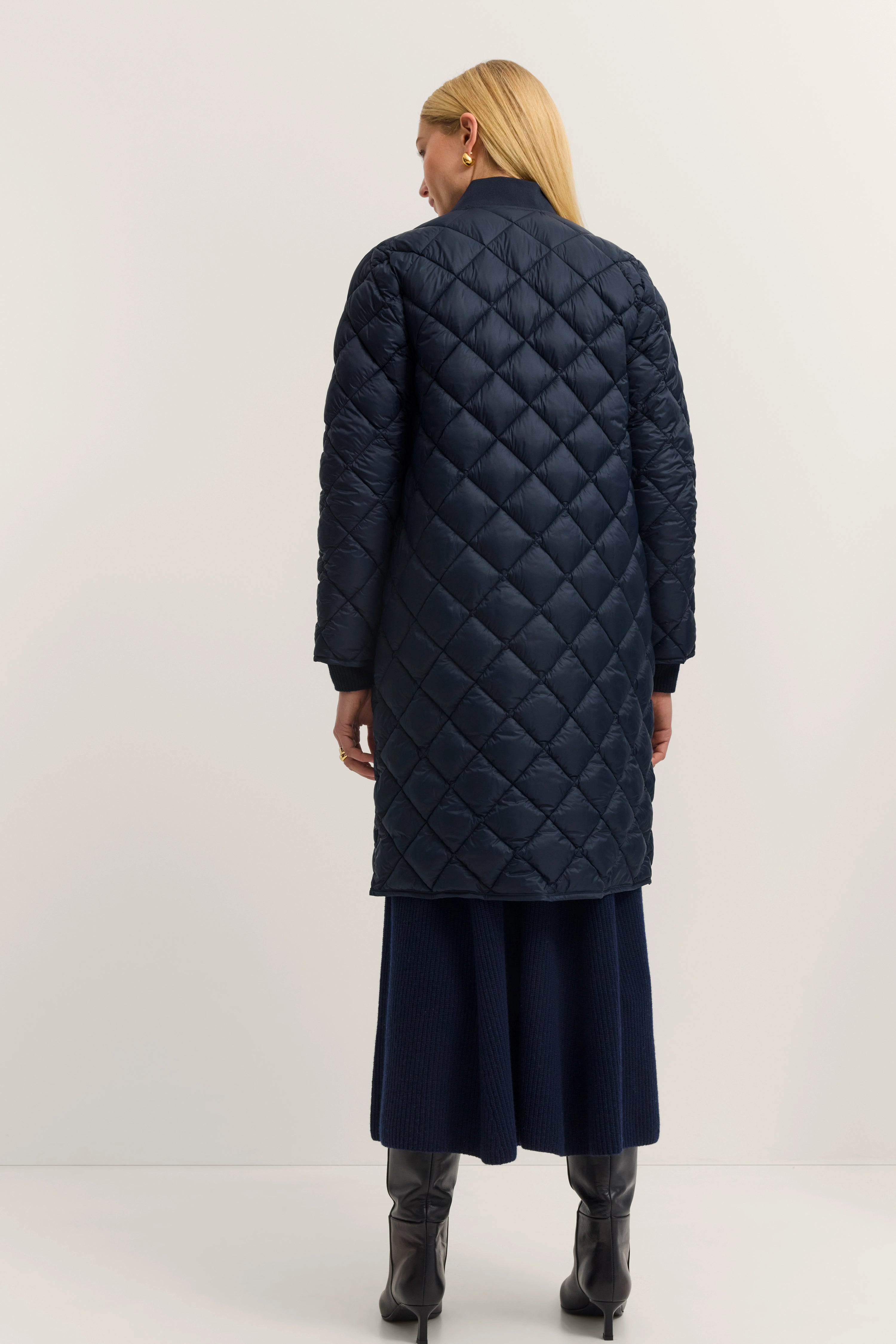 Damen-Outfit mit gesteppter, navyfarbener Mantel von bugatti, dunkelblauer Rock, schwarze Stiefel. Model von hinten.