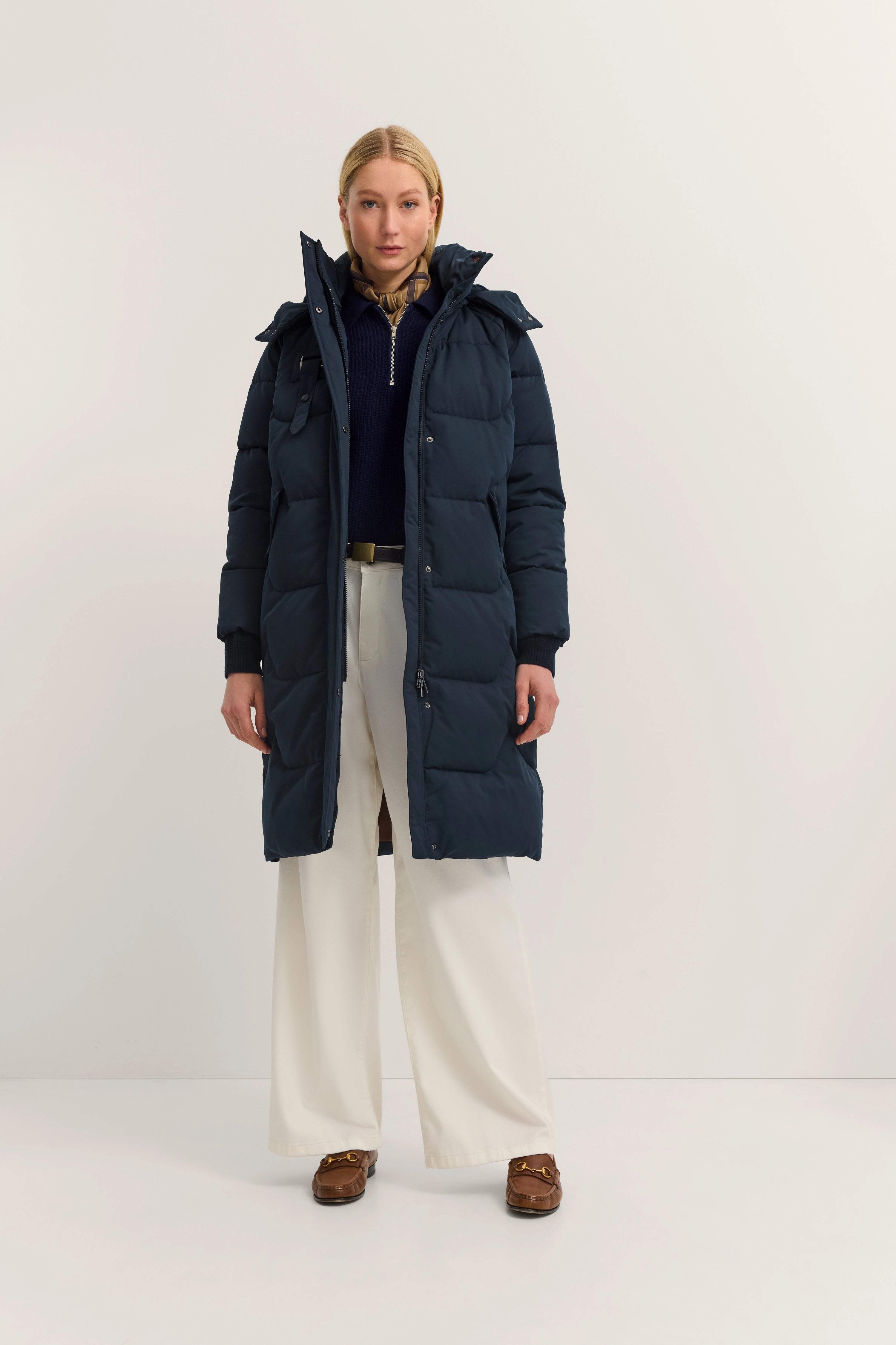 Langes, marineblaues bugatti-Daunenmantel, cremefarbene Hose, braune Schuhe, Model stehend.