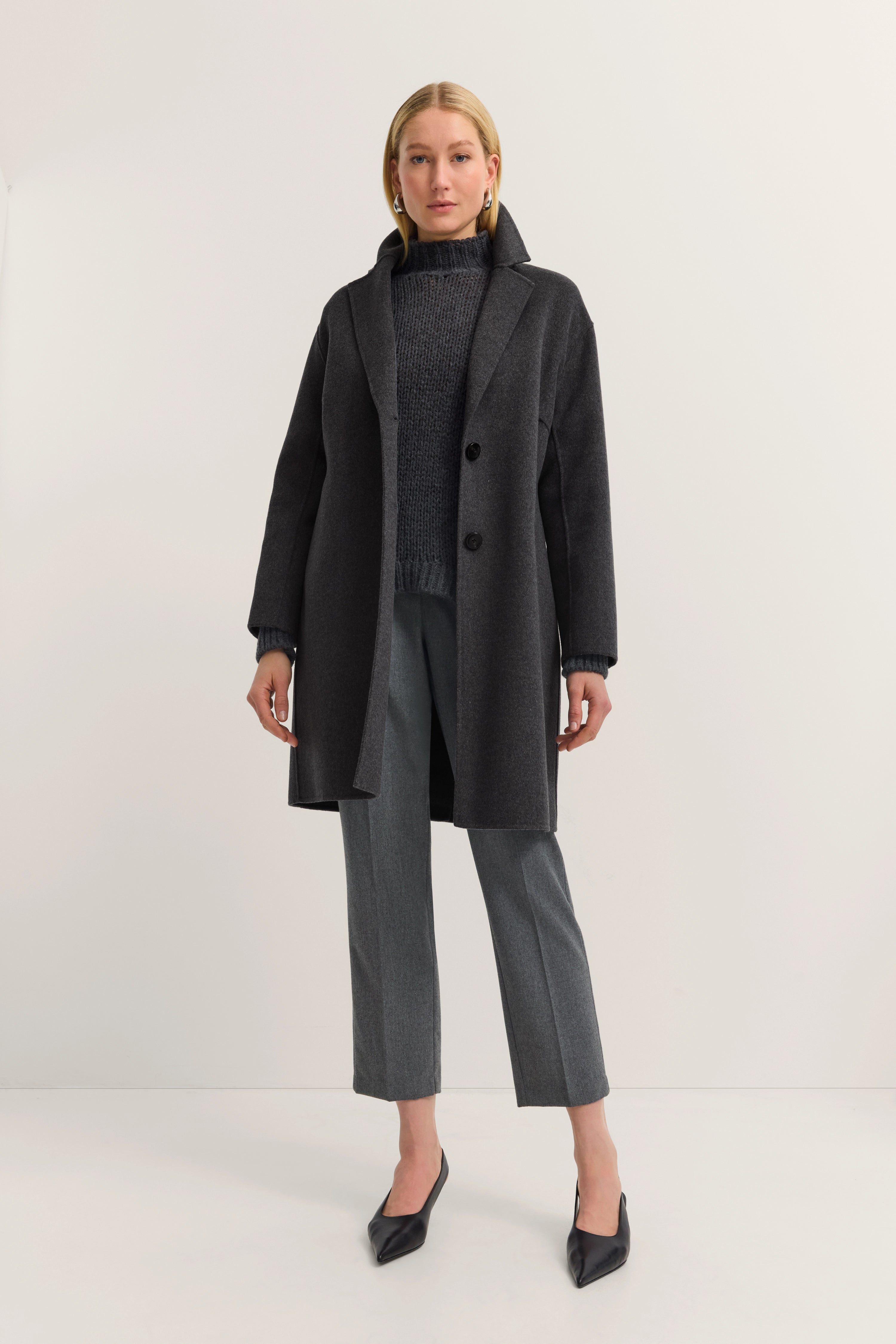 Vrouw in grijze bugatti-jas, grijze trui en broek, modelbeeld met ontspannen pose.