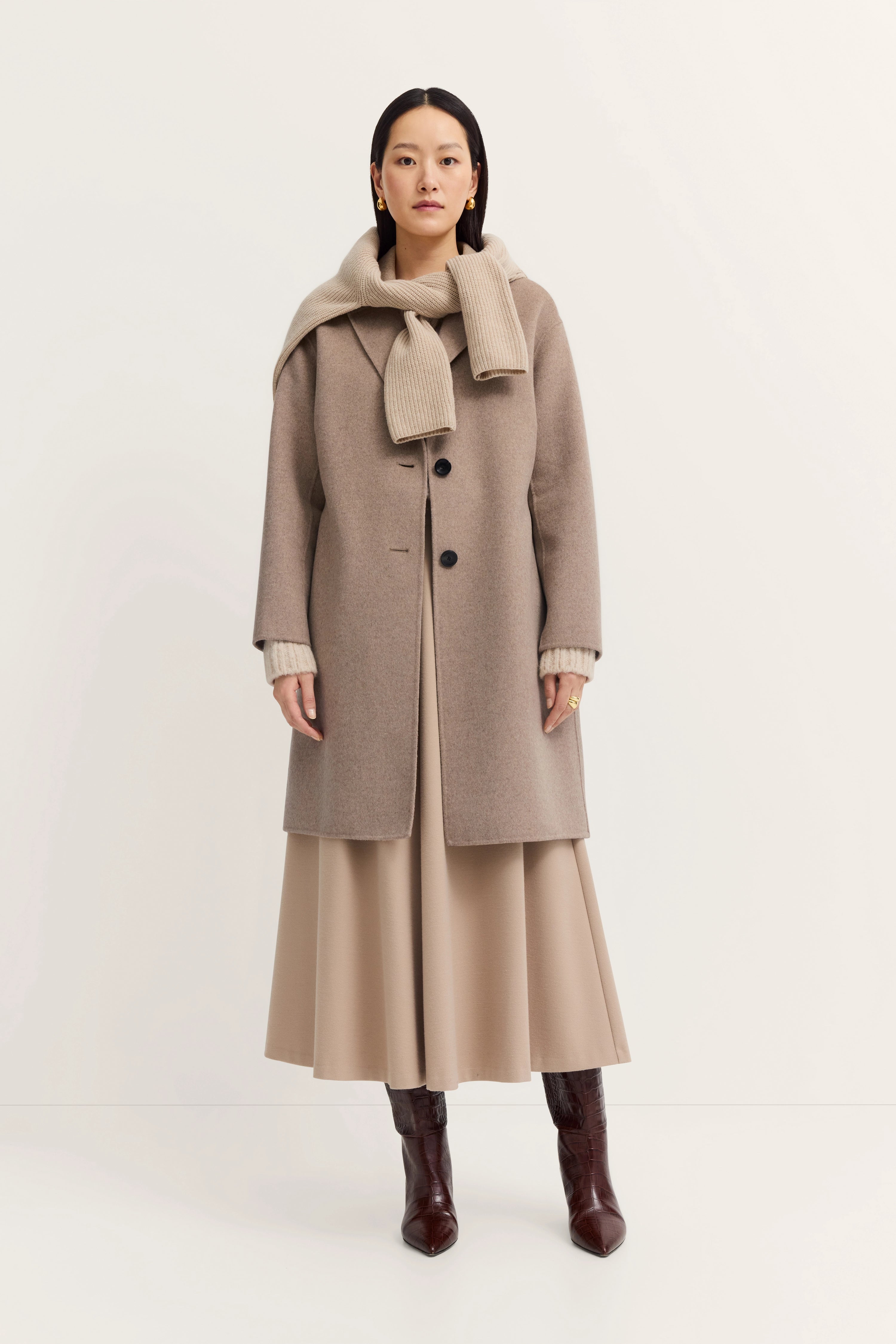 Beige bugatti mantel, trui om schouders, lange rok, bruine laarzen, model in ontspannen pose.