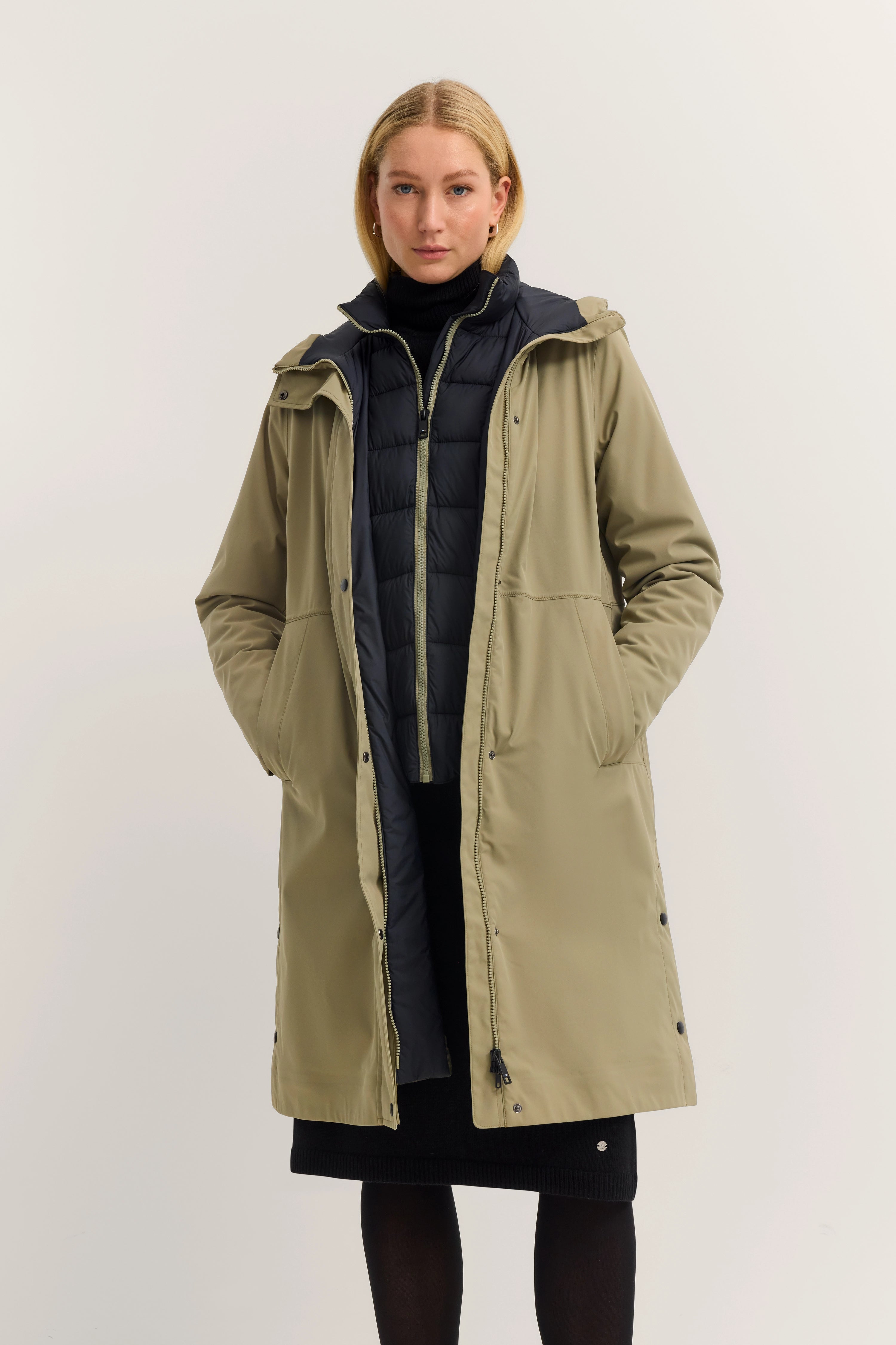 Model in olivgrünem bugatti Mantel über schwarzer Jacke, frontal stehend, Hände in Taschen.