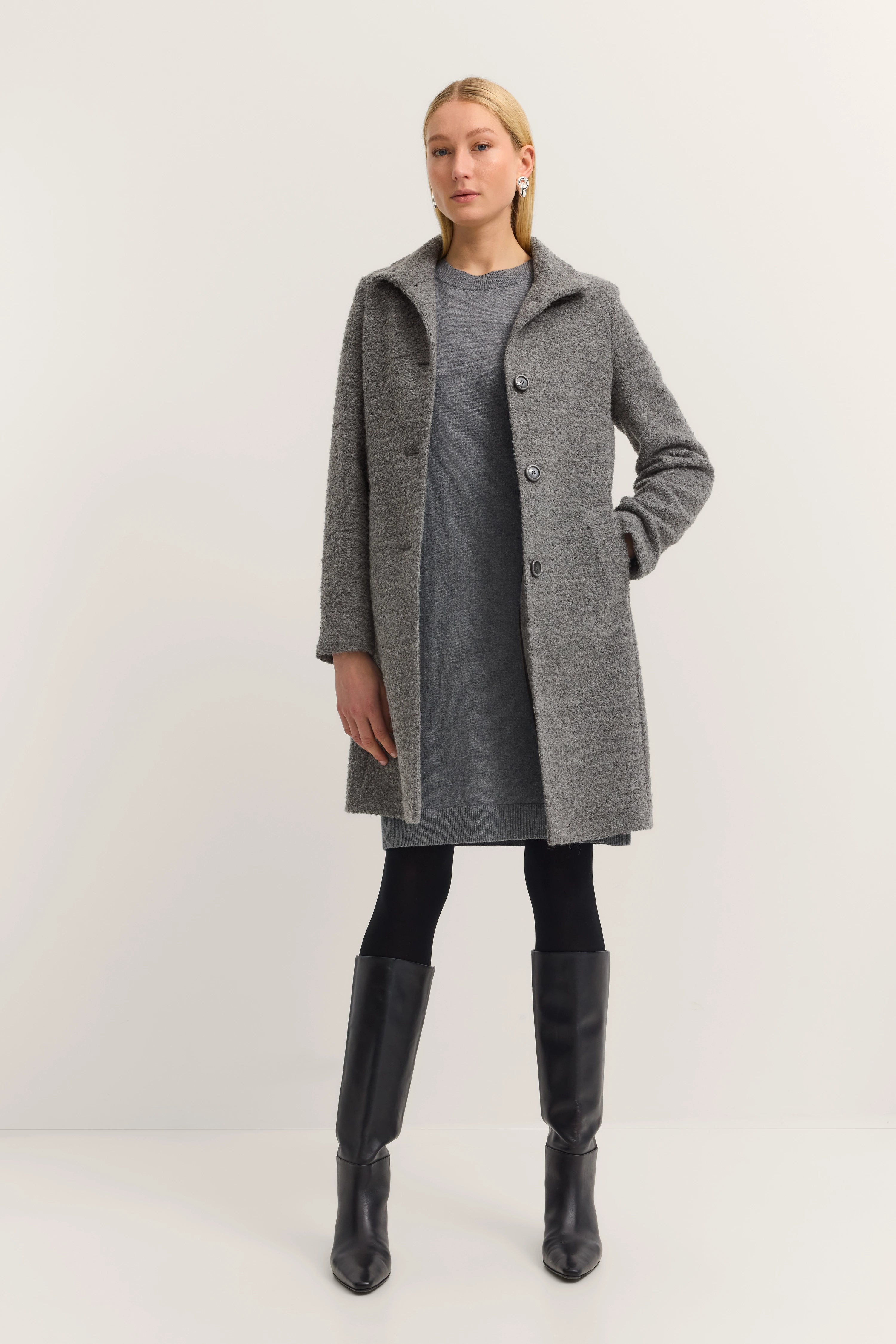 Grauer bugatti Mantel, graues Kleid, schwarze Stiefel. Model stehend mit Hand in Tasche.