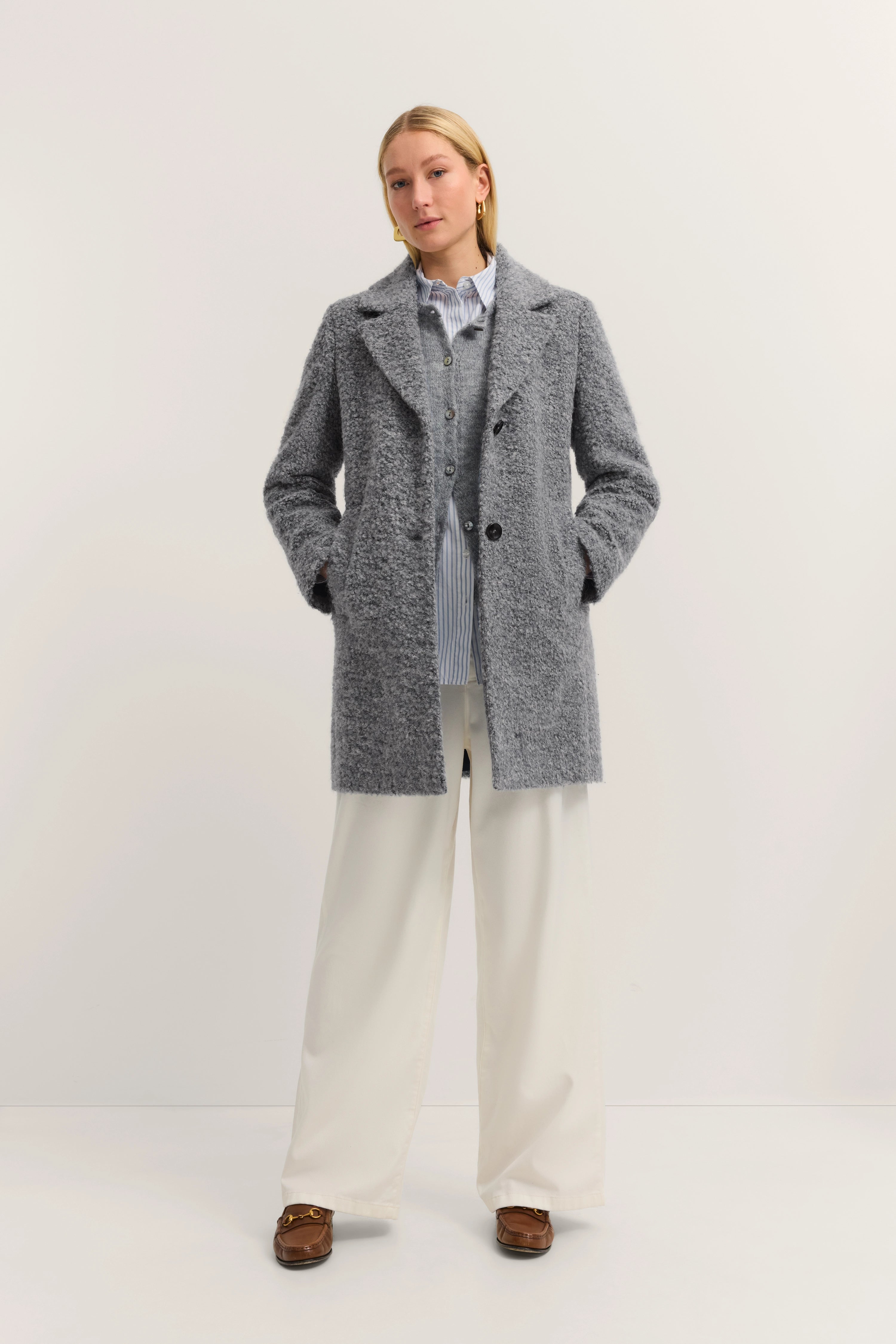 Grauer Mantel, hellblaue Bluse, weiße Hose, braune Schuhe, Model mit Händen in Taschen, frontal stehend.