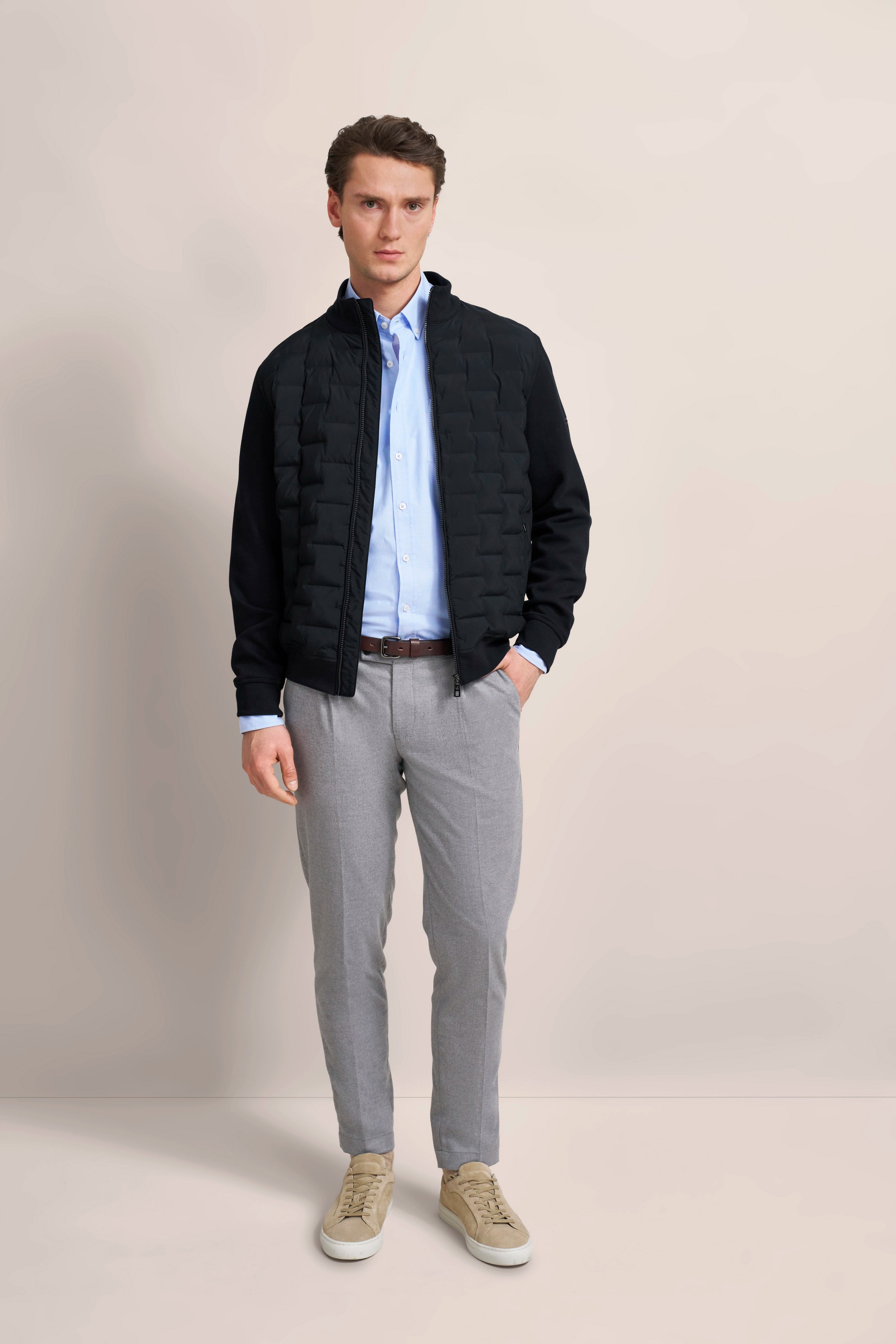 Model in schwarzer Jacke, blauem Hemd, grauer Hose, beigem Sneaker. Lässig stehend, Hände in Taschen.