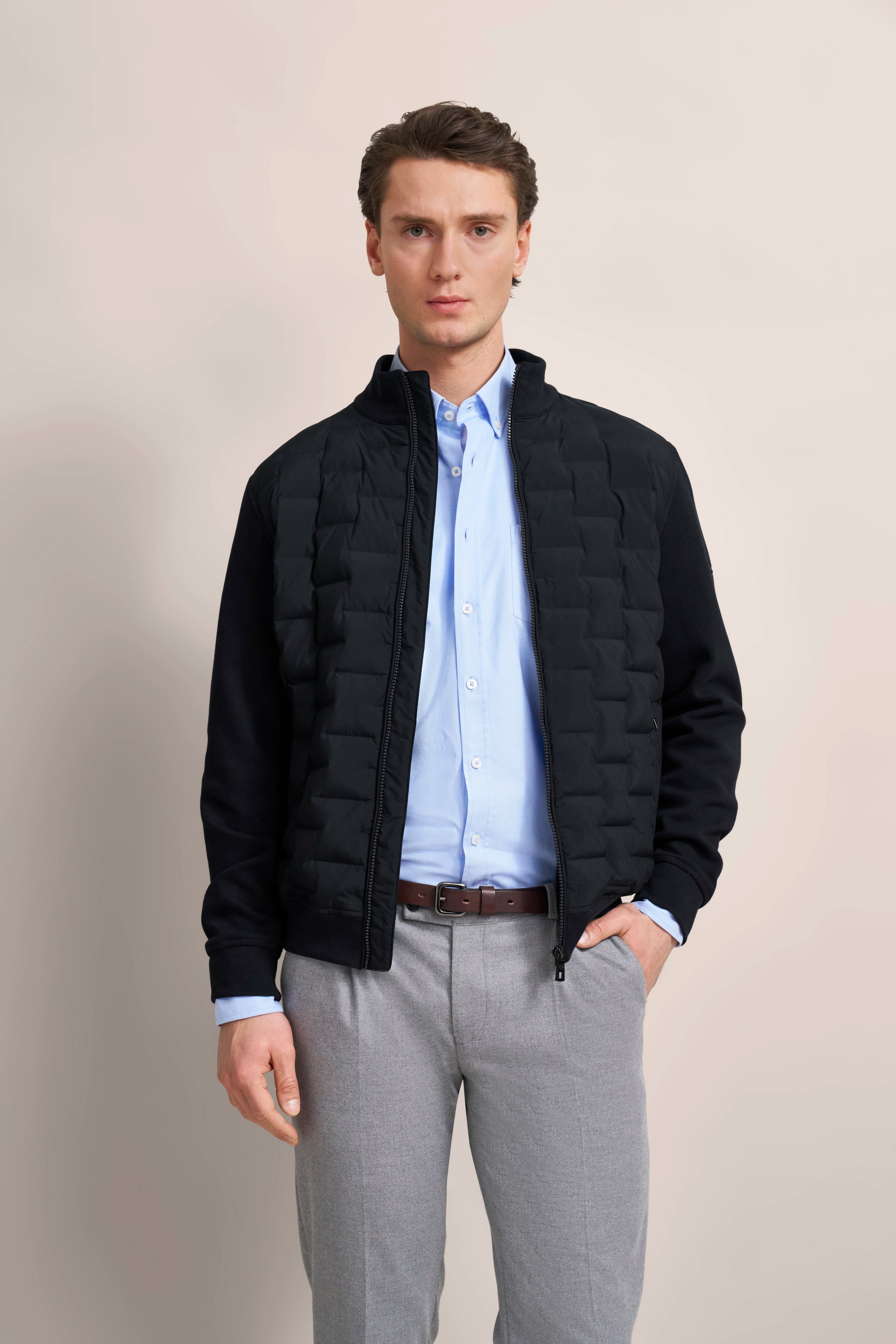 bugatti Model in schwarzer Steppjacke, hellblauem Hemd, grauer Hose, entspanntes Posing.