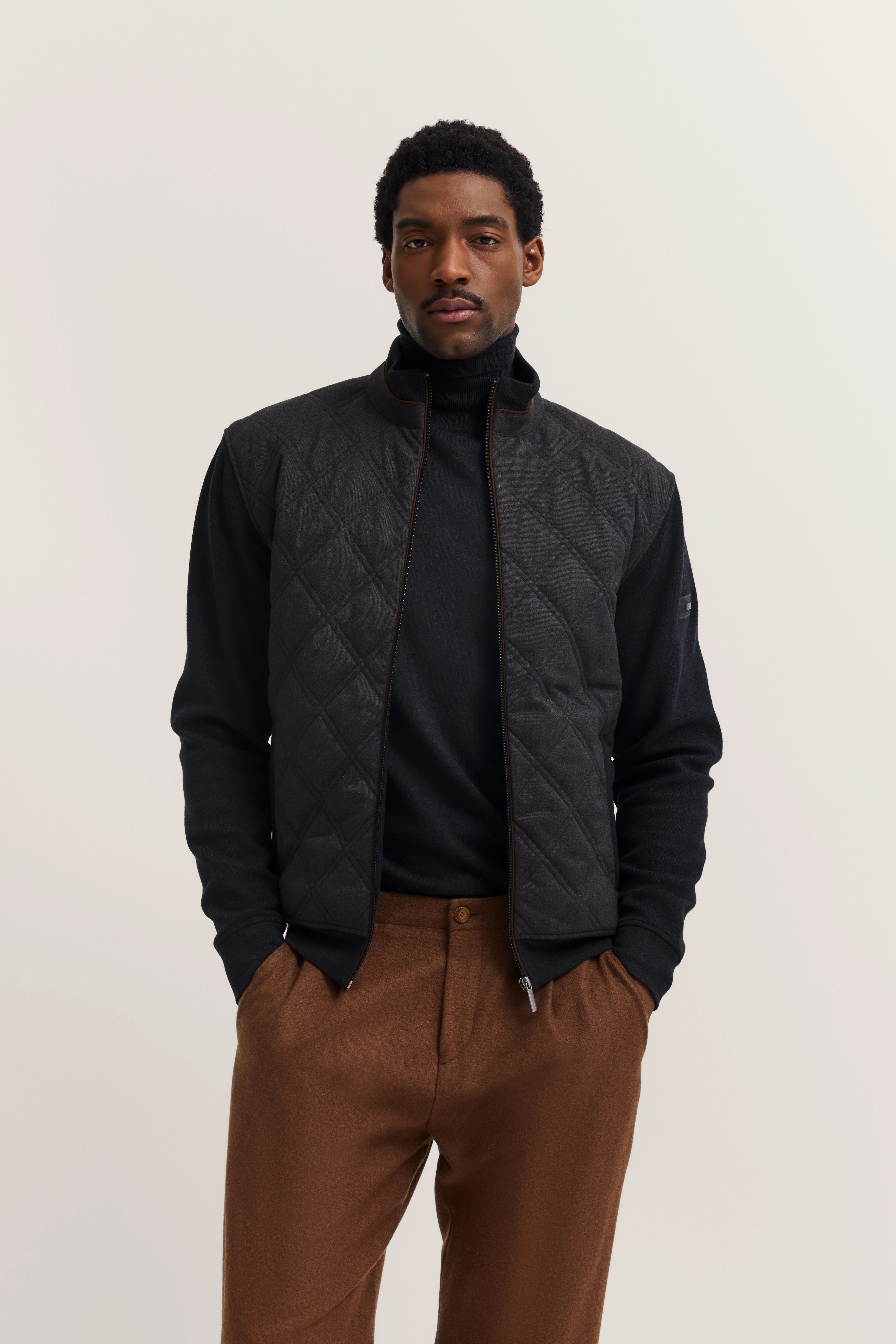 Schwarze Steppjacke, schwarzer Pullover, braune Hose, Model steht lässig, bugatti Stil.