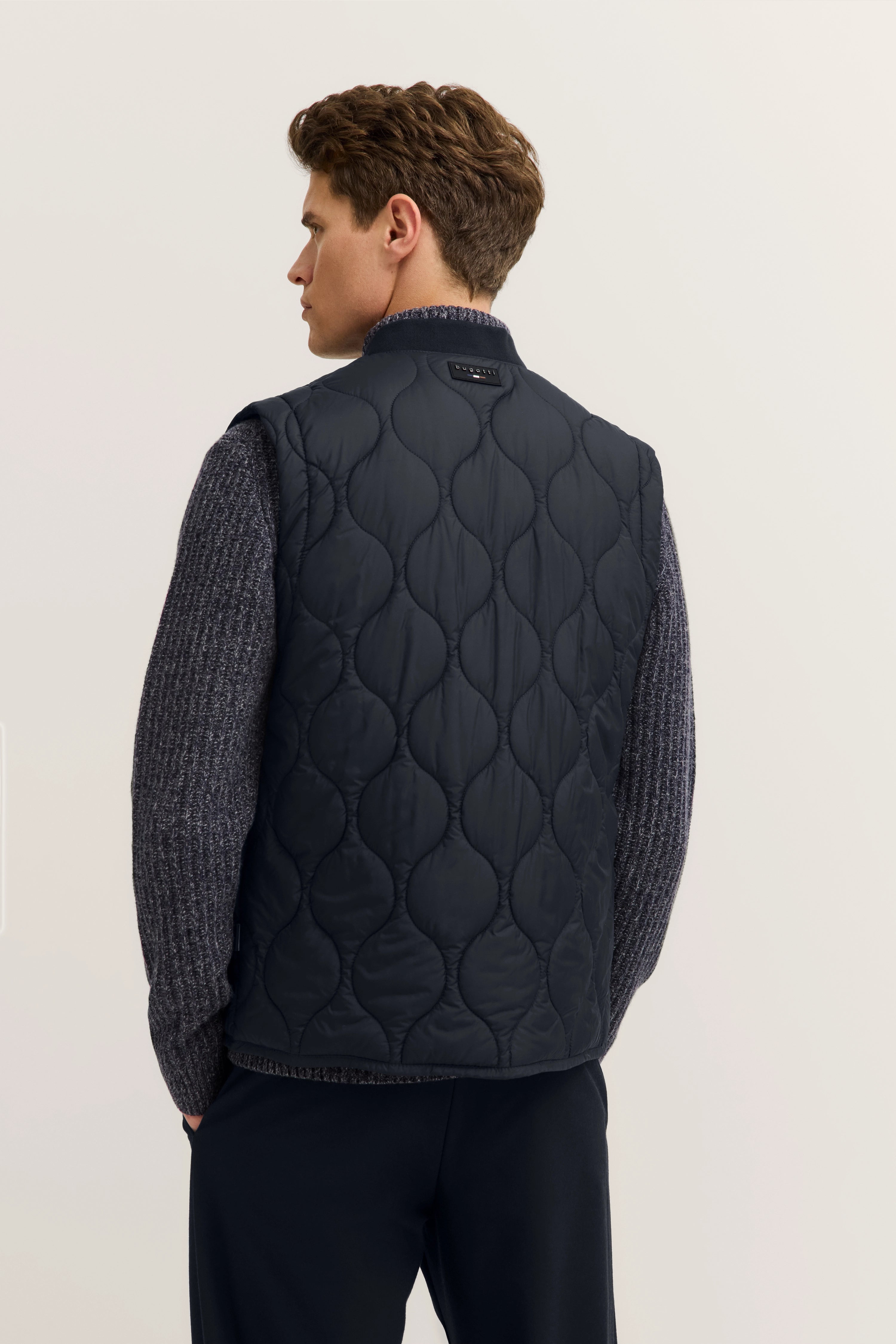 Rückansicht eines Models in dunkelblauer Steppweste und grauem Strickpullover von bugatti.