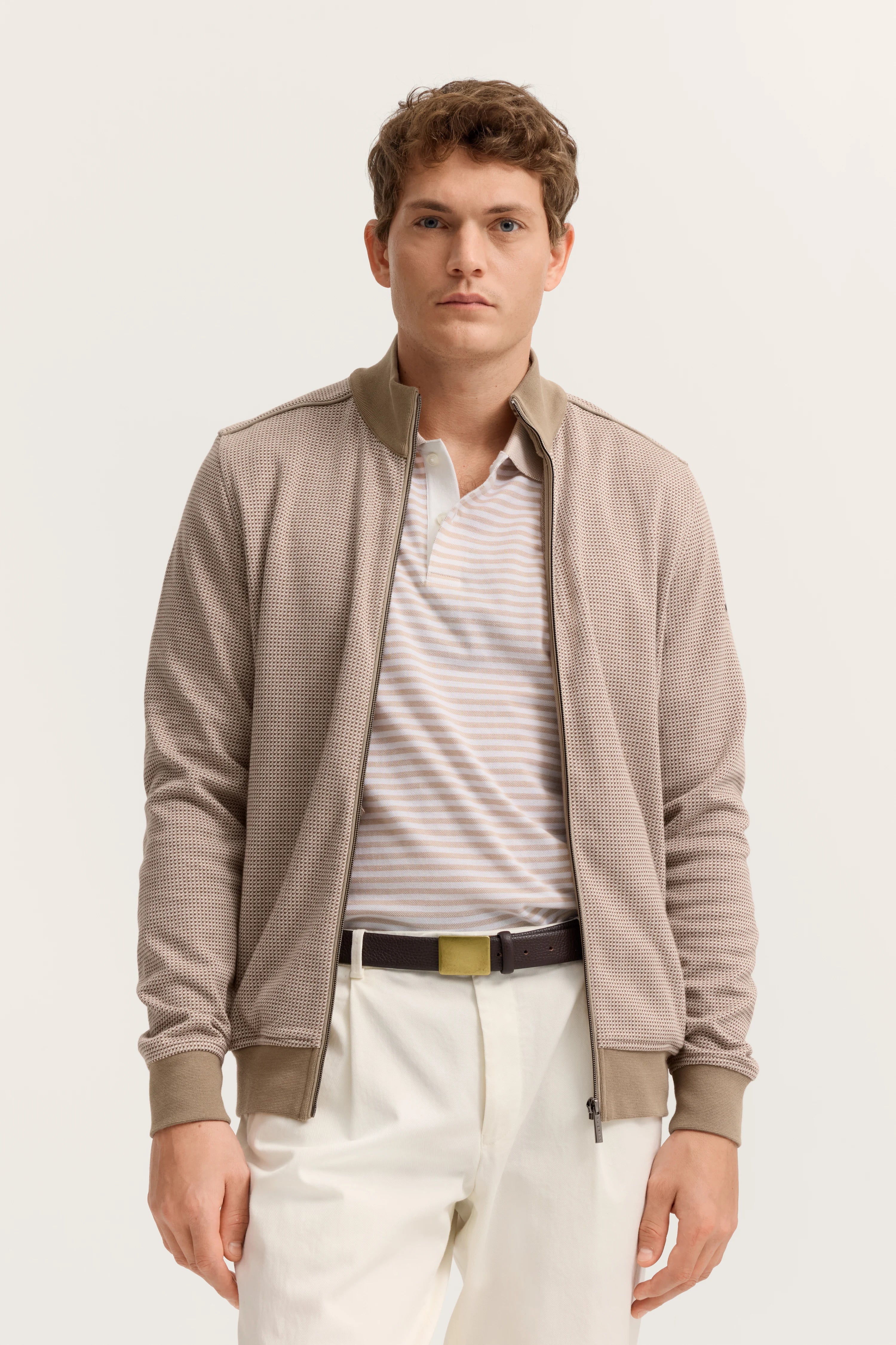 Beiges bugatti Model-Outfit mit gestreiftem Polo, beige Jacke und weißer Hose, entspanntes Posing.