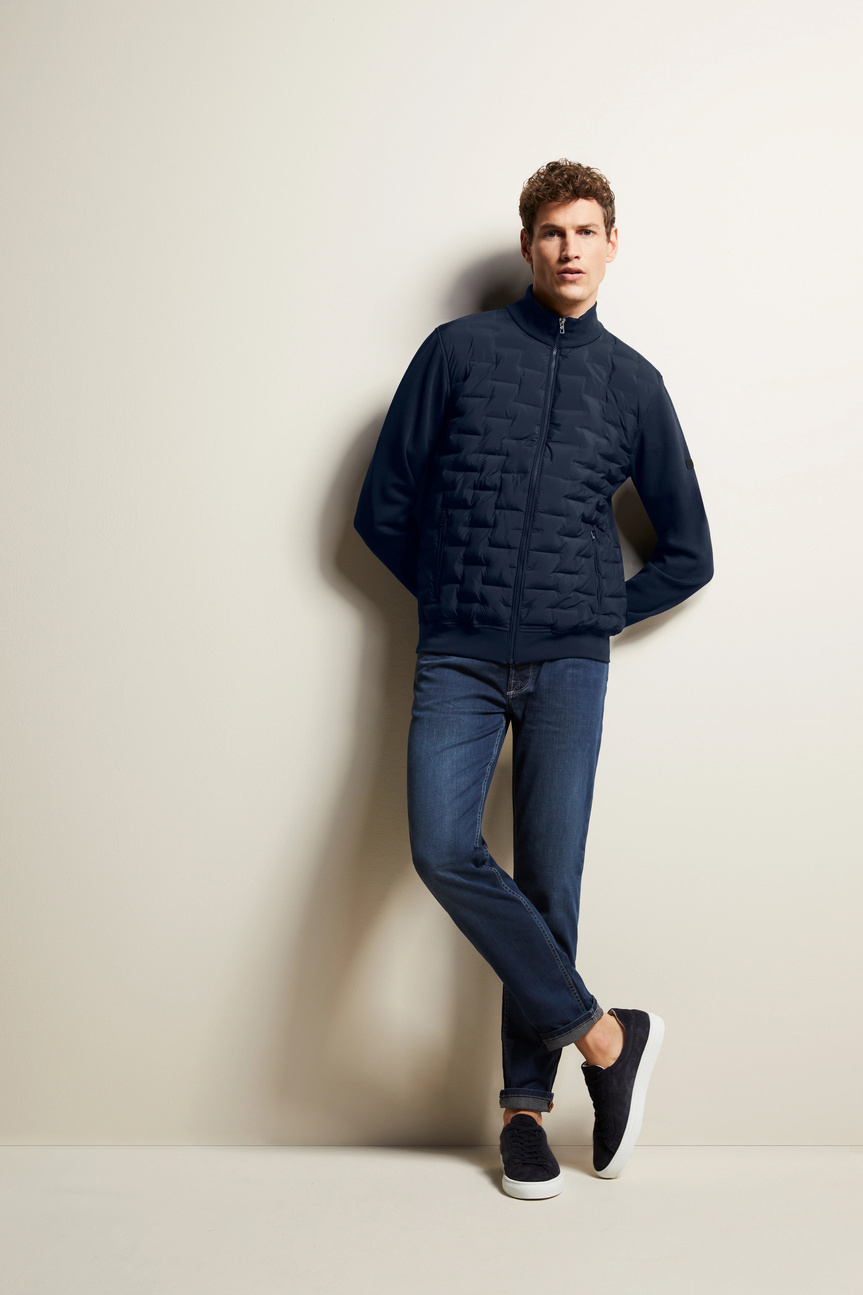 Model trägt blaue bugatti Steppjacke, Jeans und Sneakers. Lässige Pose, an Wand gelehnt.