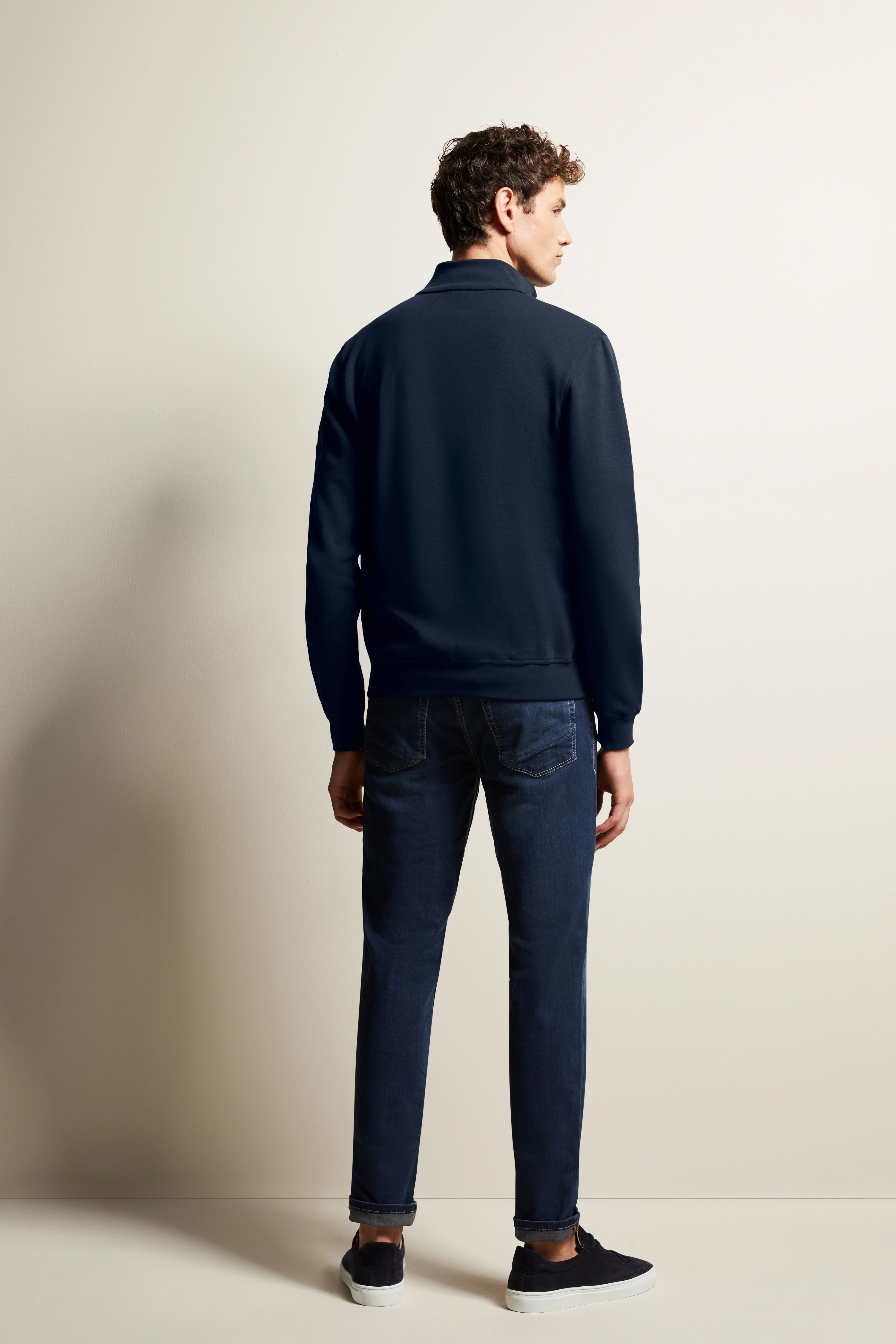 Model trägt dunkelblaue bugatti Jacke, Jeans und Sneakers, Rückansicht.