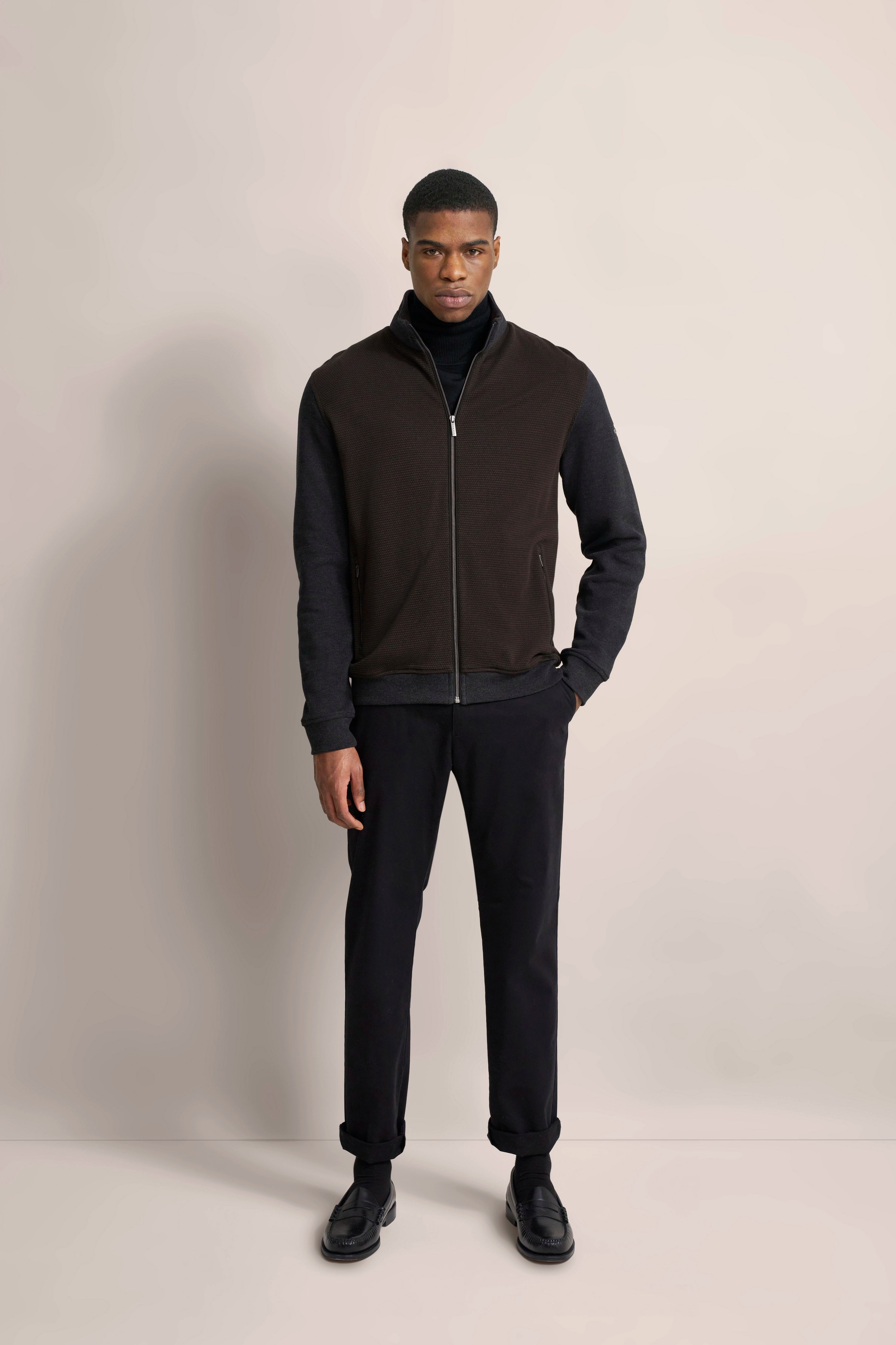"bugatti Outfit: Braune Weste, schwarzer Pullover, schwarze Hose, Model stehend, entspanntes Posing."