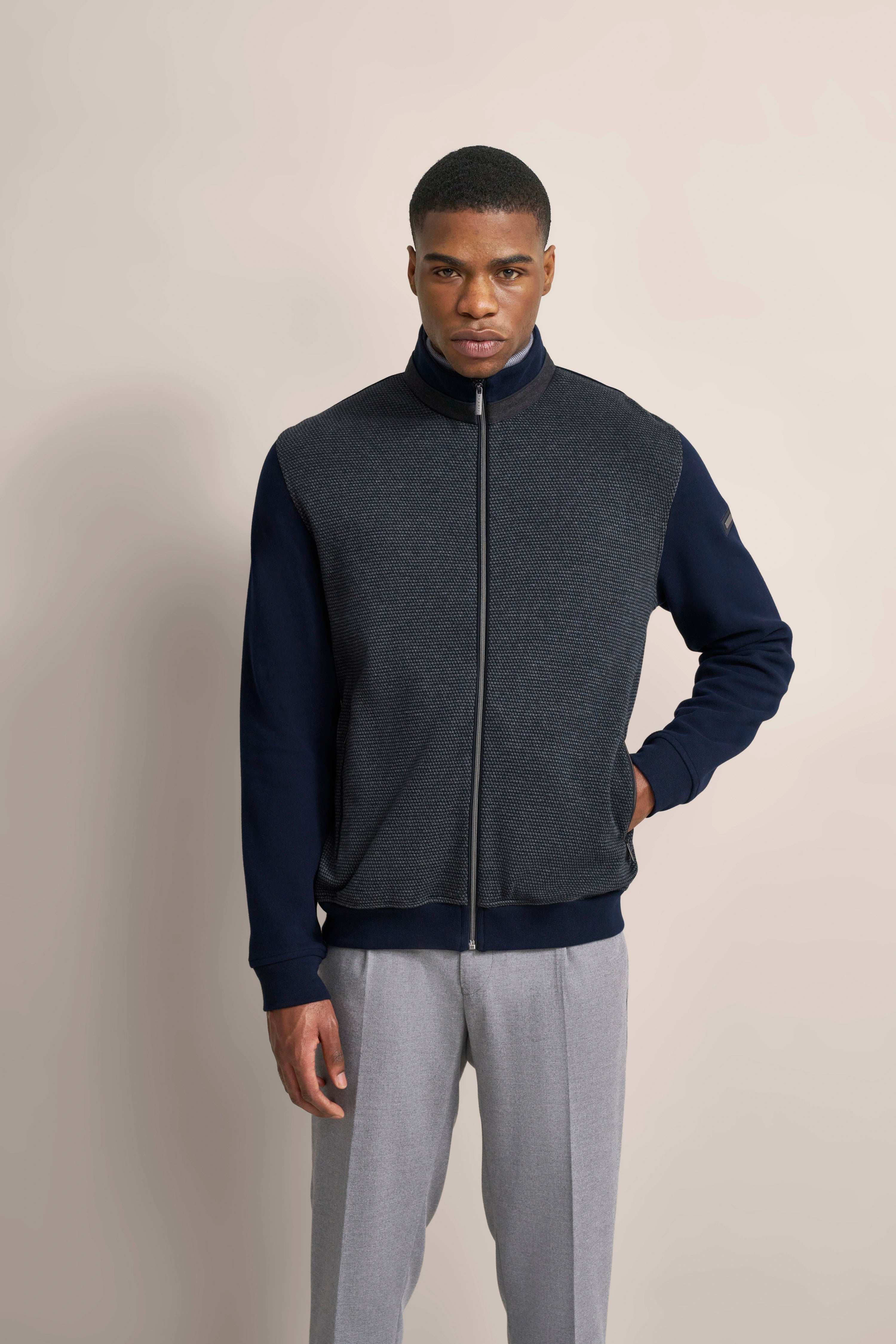 Model in grauer Weste und blauer Jacke, graue Hose, neutraler Hintergrund, gerade Haltung.