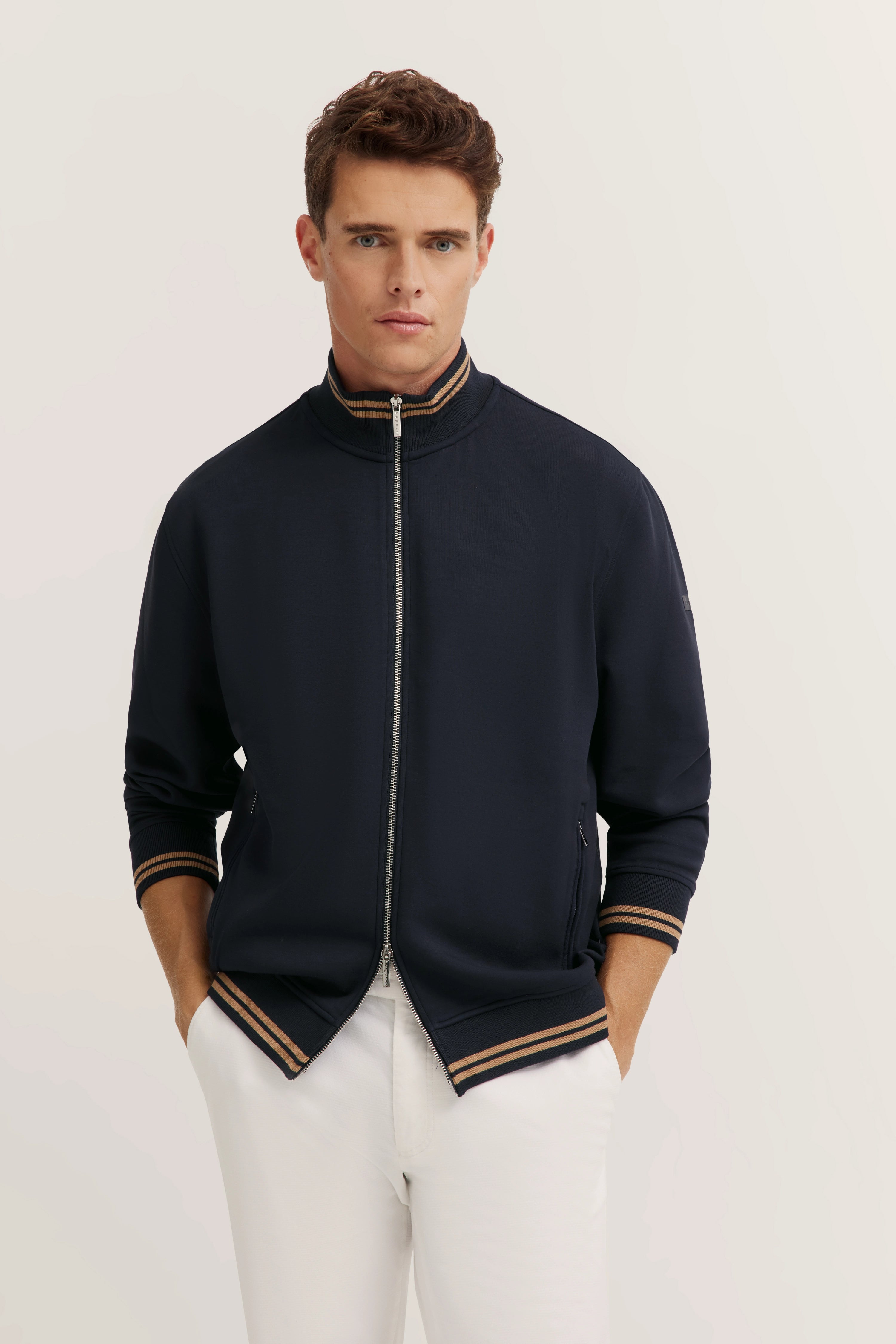 bugatti Model in navy Jacke mit braunen Streifen, weißer Hose, entspannte Pose, zeitloser Stil.