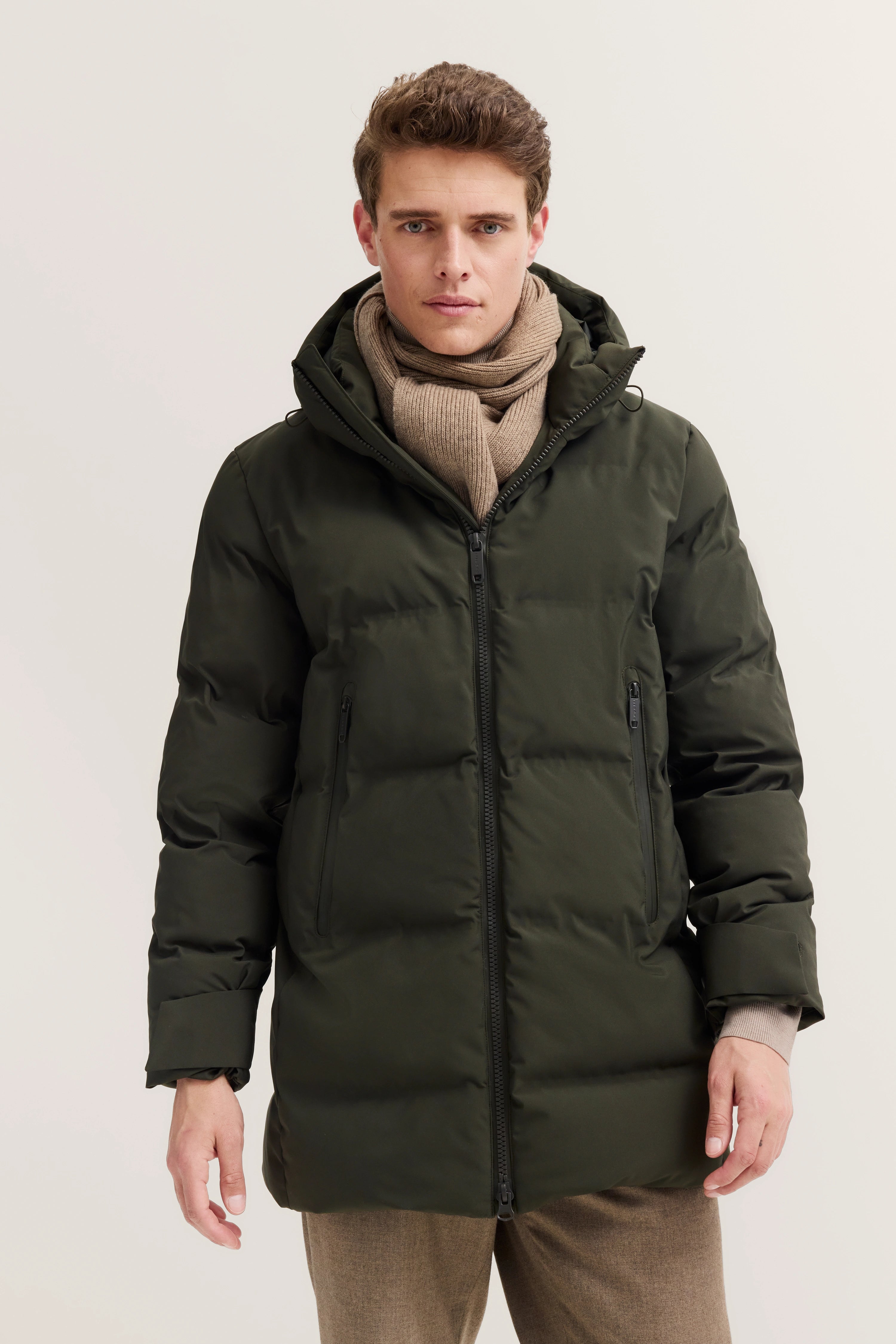 Dunkelgrüne bugatti Daunenjacke und beigefarbener Schal, Modelbild, entspanntes, selbstbewusstes Posing.